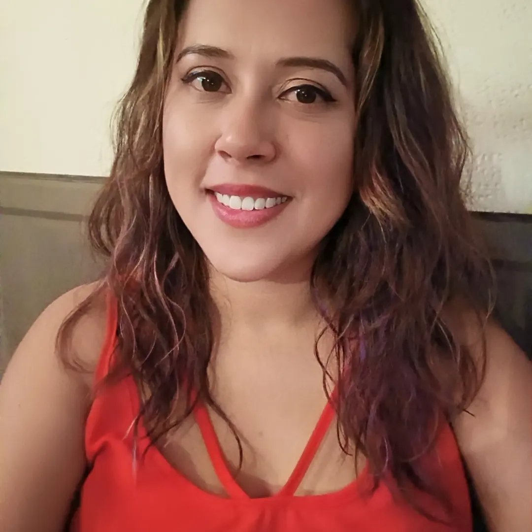 Woman with wavy brown hair and a red sleeveless top smiles at the camera in a casual setting.