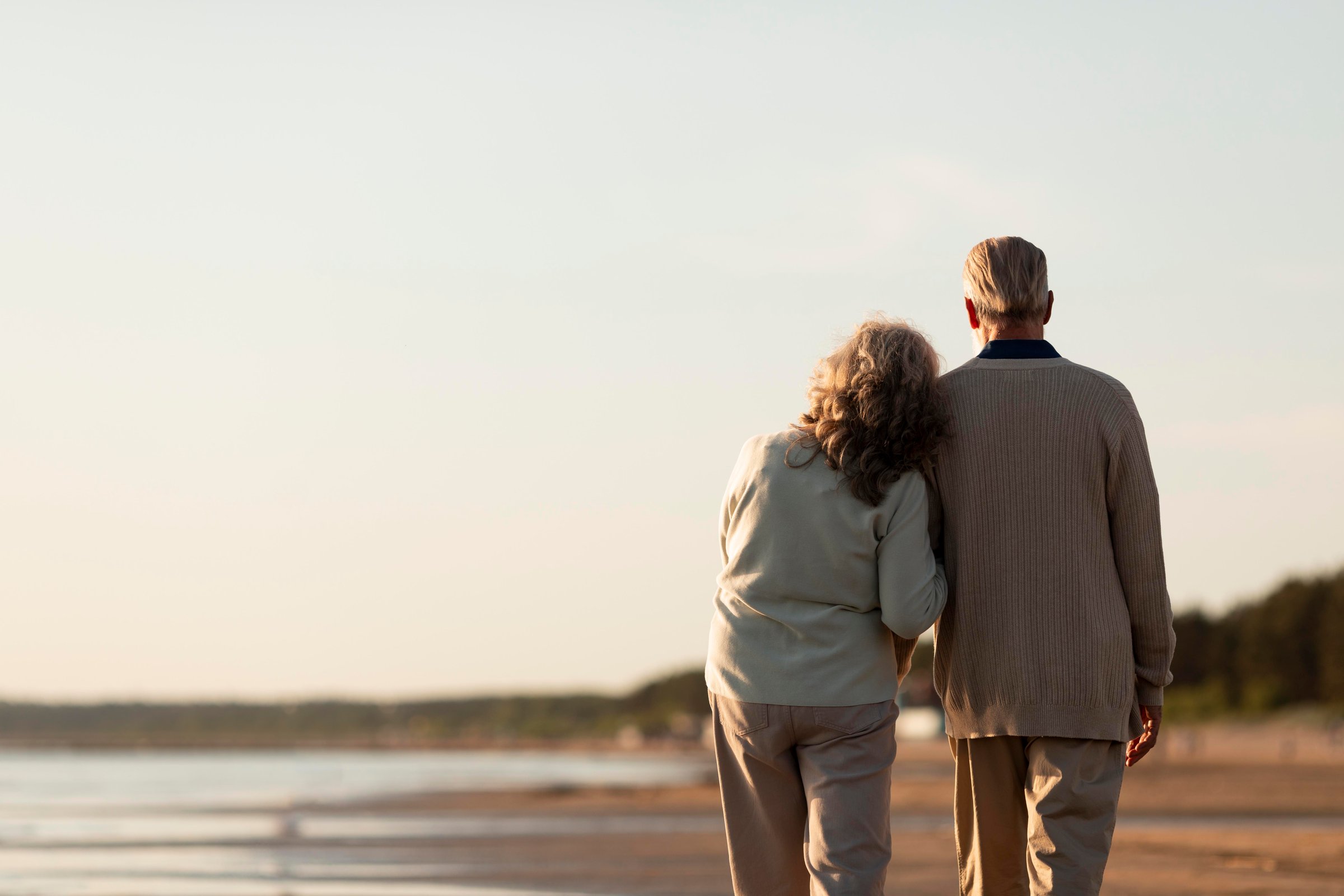 Elderly couple walking arm in arm along a serene beach, gazing at the water, with a clear sky and distant trees.