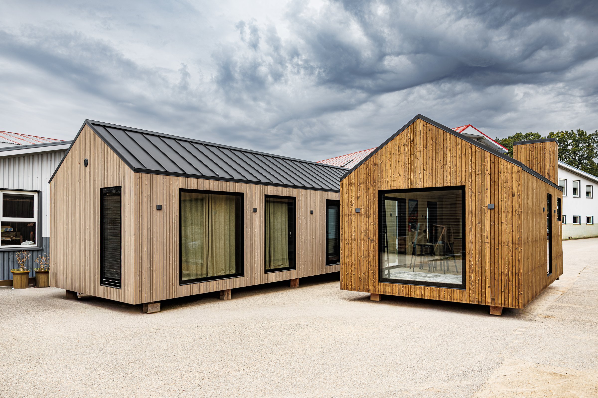 Two modern wooden tiny houses with vertical cladding, dark metal gable roofs, and elevated bases, showcasing sustainable modular architecture under a cloudy sky