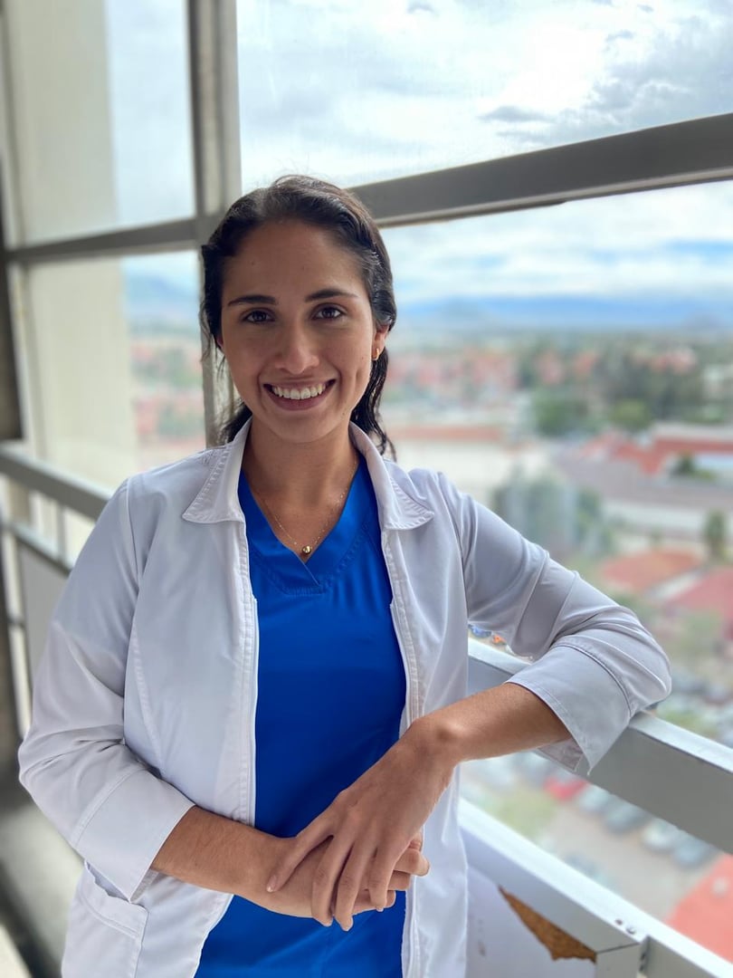 Smiling woman in blue scrubs and white coat standing by a window with a city view in the background.