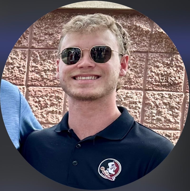 Young man in sunglasses, wearing a black polo with a logo, standing against a brick wall, smiling at the camera.