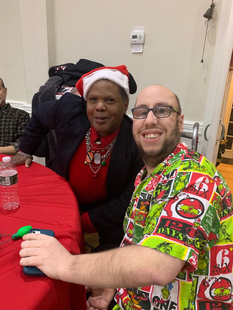 Two people smiling at a festive gathering; one wears a Santa hat, the other a holiday-themed shirt. They're seated at a red table.
