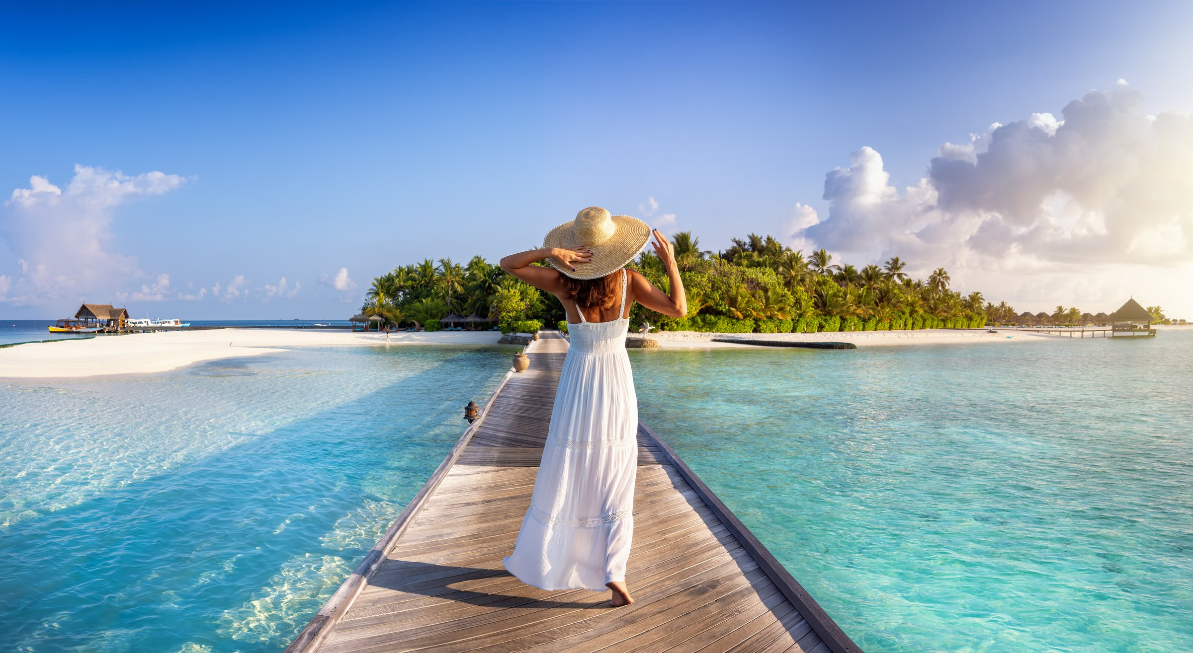 Back view of a elegant woman in white dress and hat walks down a pier towards a tropical island in the Maldives