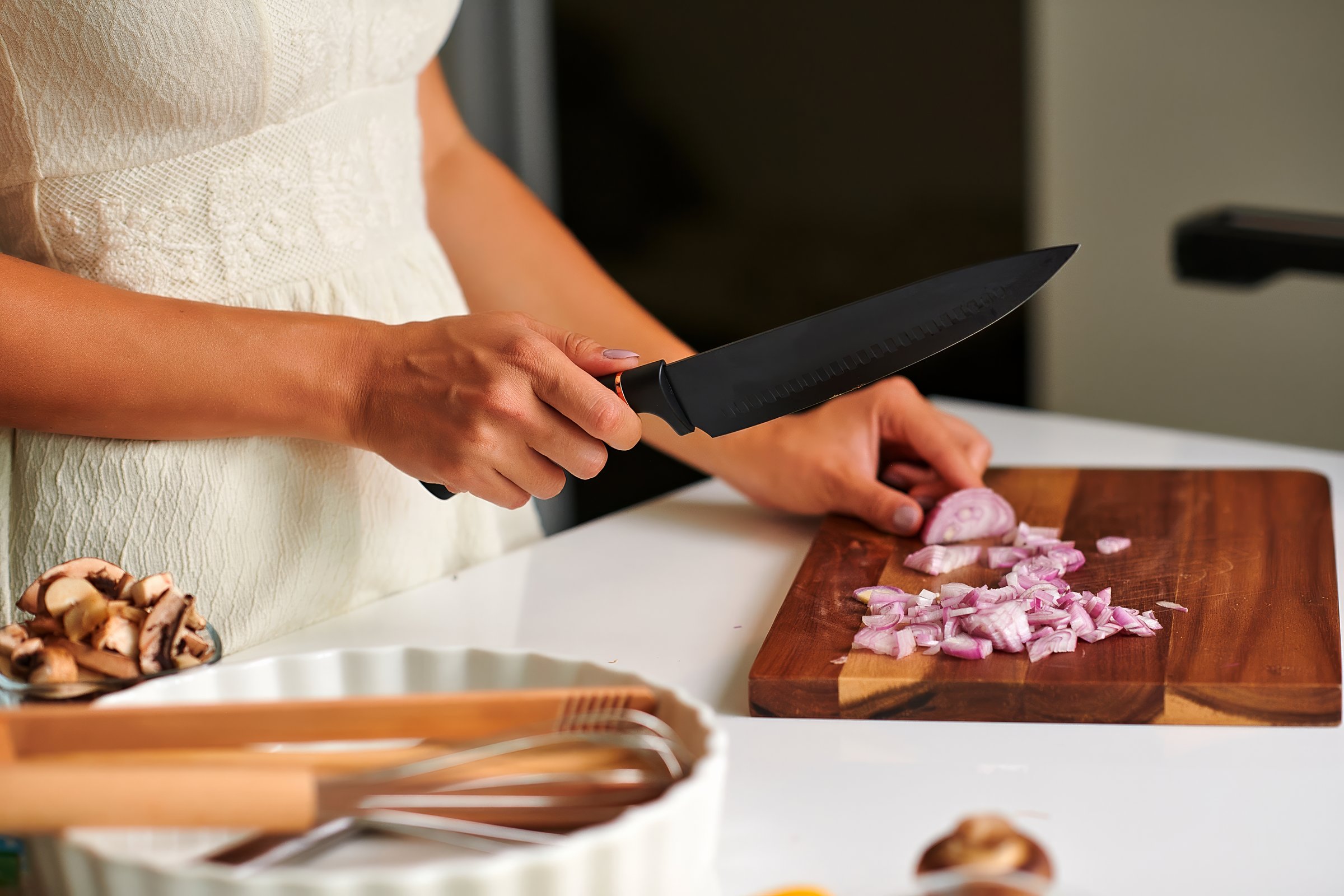 Female chef in a white dress is skillfully chopping shallots on a wooden cutting board with a knife, surrounded by fresh ingredients and kitchen tools in a bright modern kitchen atmosphere