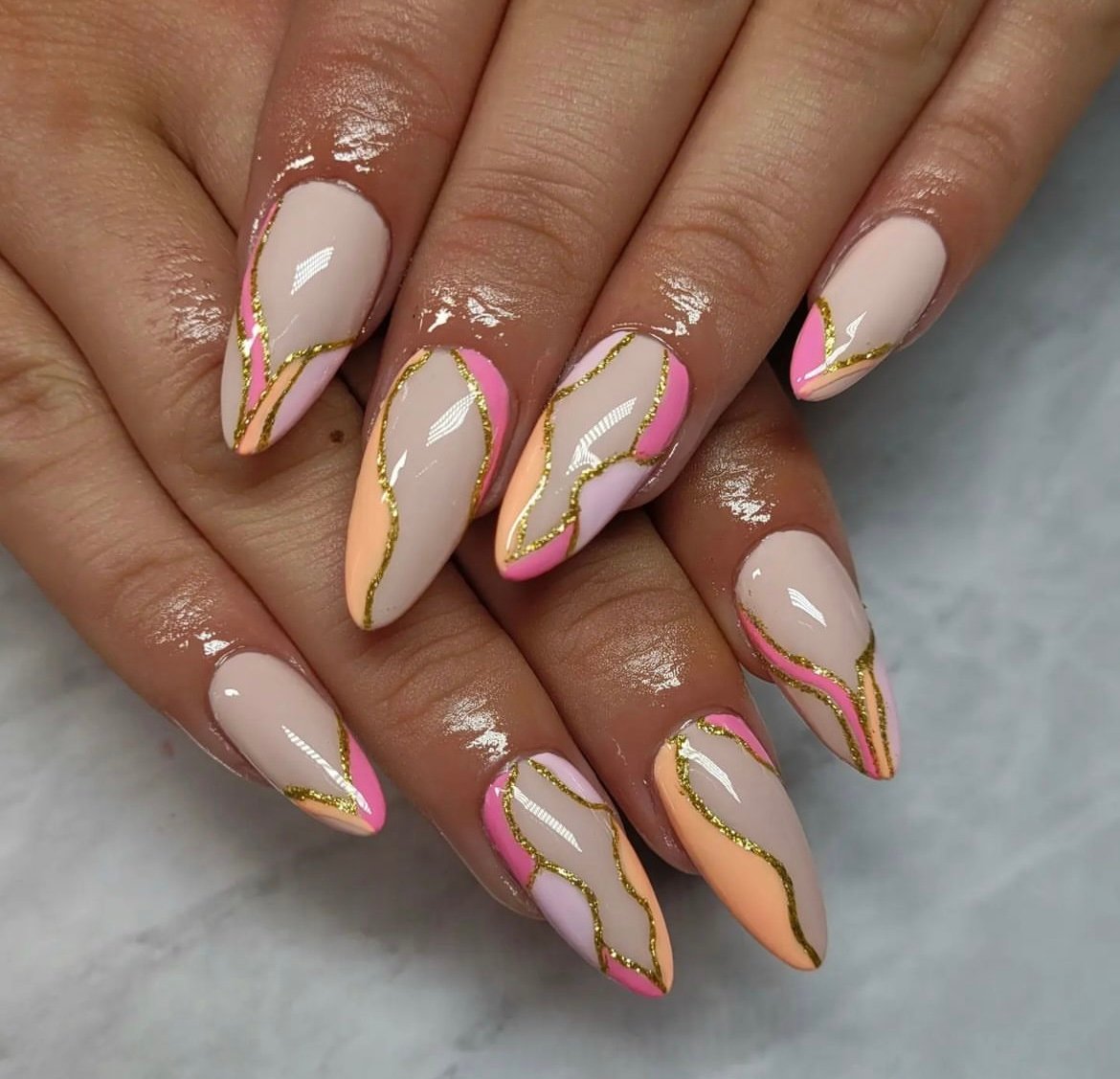 Hands with long almond-shaped nails featuring a glossy multicolored design and gold accents on a marble-like surface.