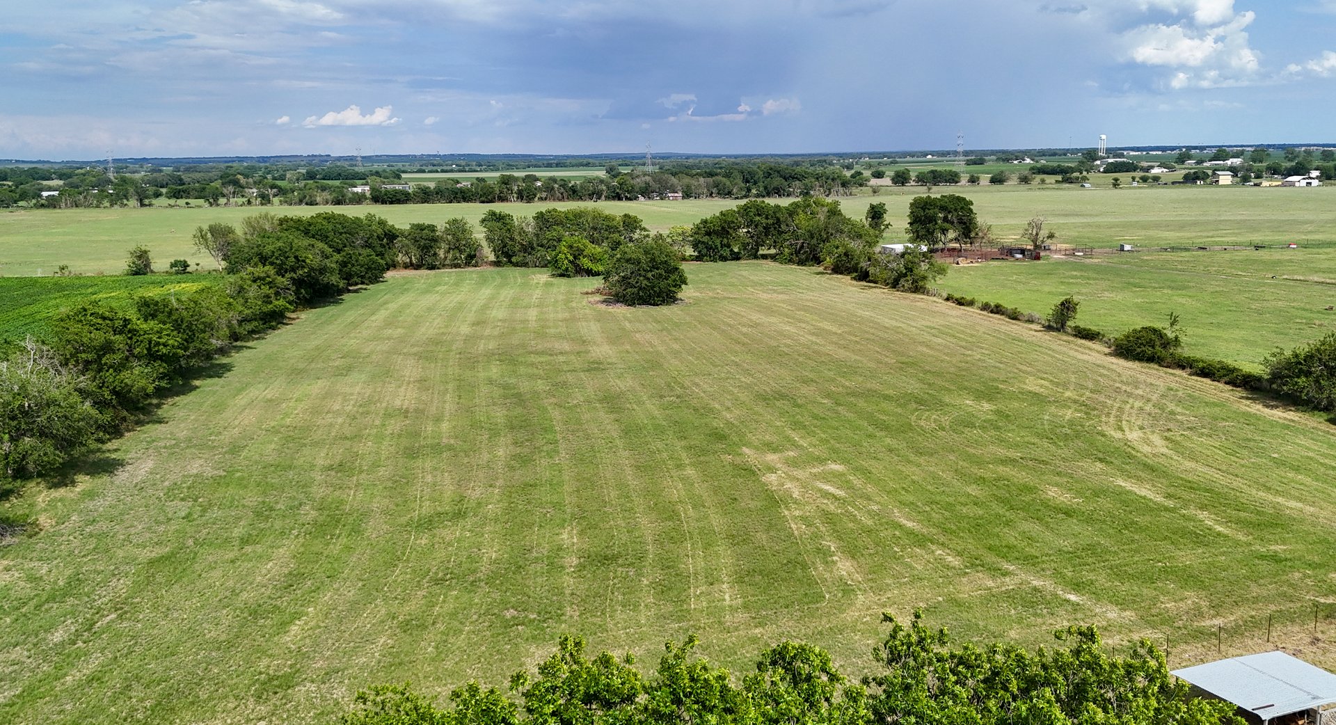 Aerial view of a large, open green field surrounded by trees under a partly cloudy sky.