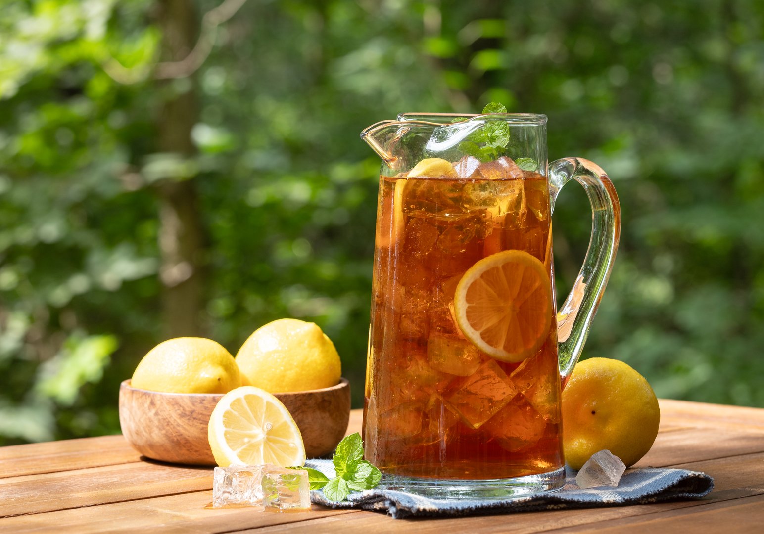 Pitcher of ice tea with lemon slices and mint outdoors on wooden table with nature background