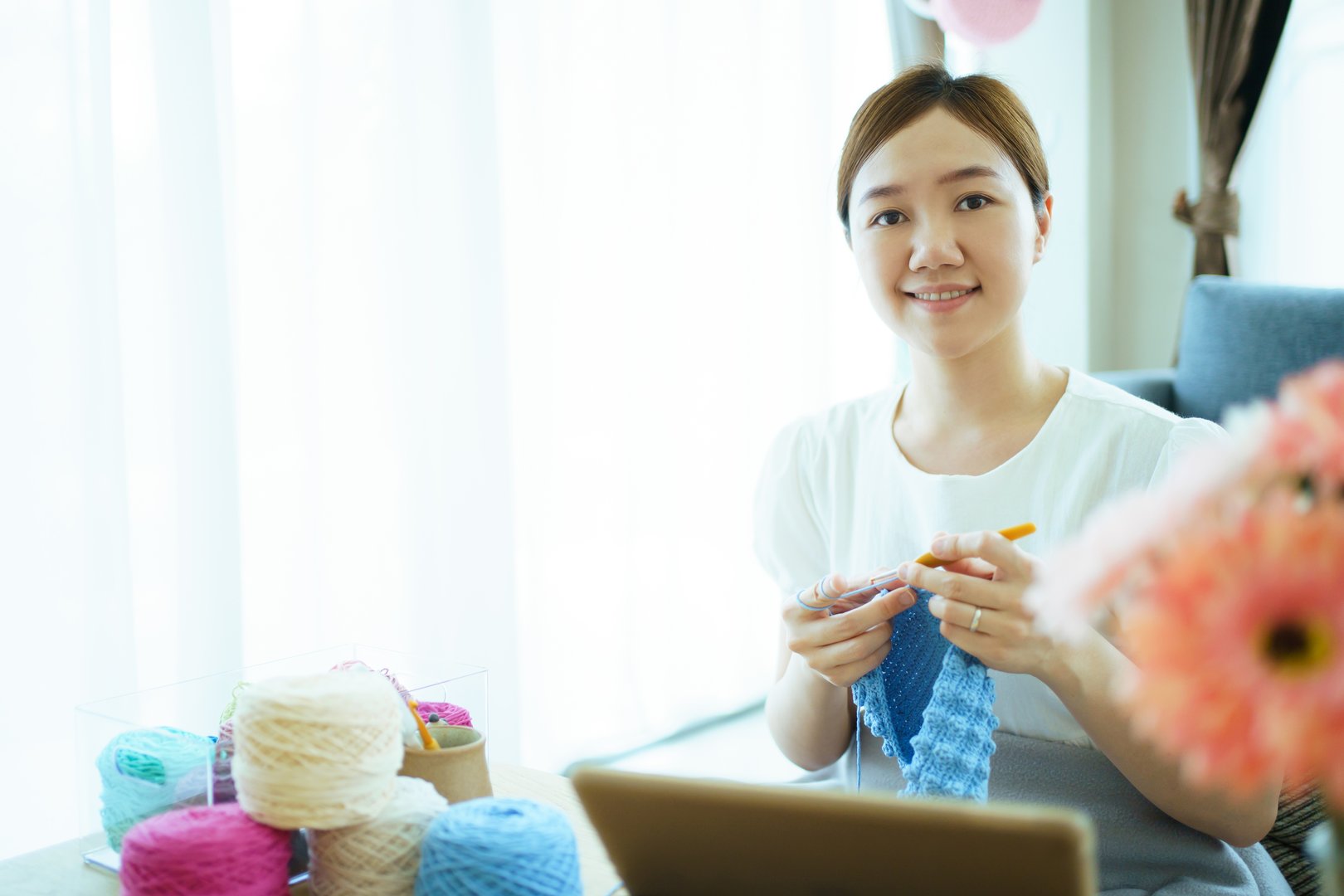 Happy cheerful Asian woman doing a crochet in living room in free time. Skillful Asian craftswoman knitting a handcraft crochet sweater and hat in cozy living room.