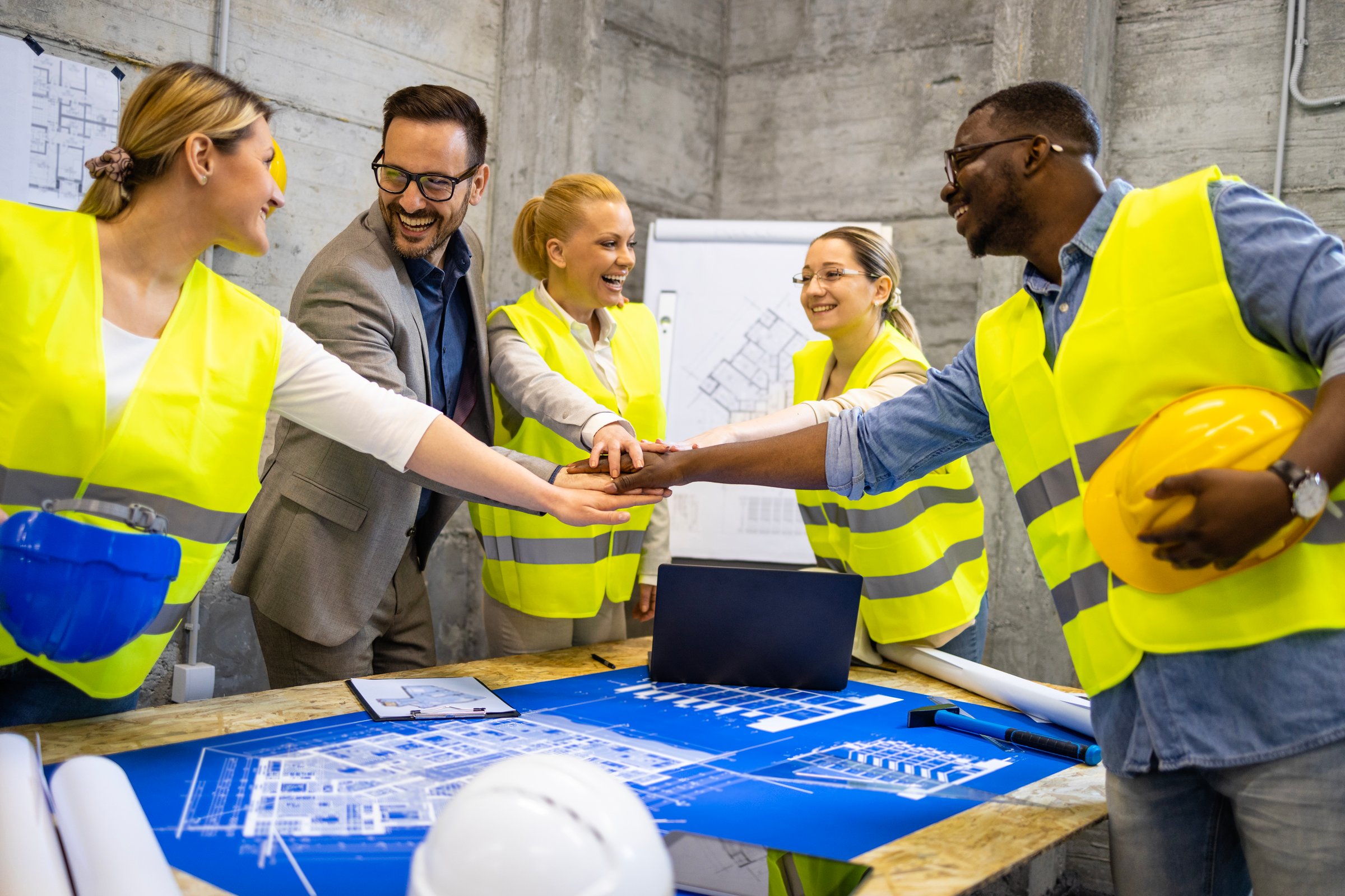 Team of structural engineers and architects holding hands together above building plans and blueprints at construction site.
