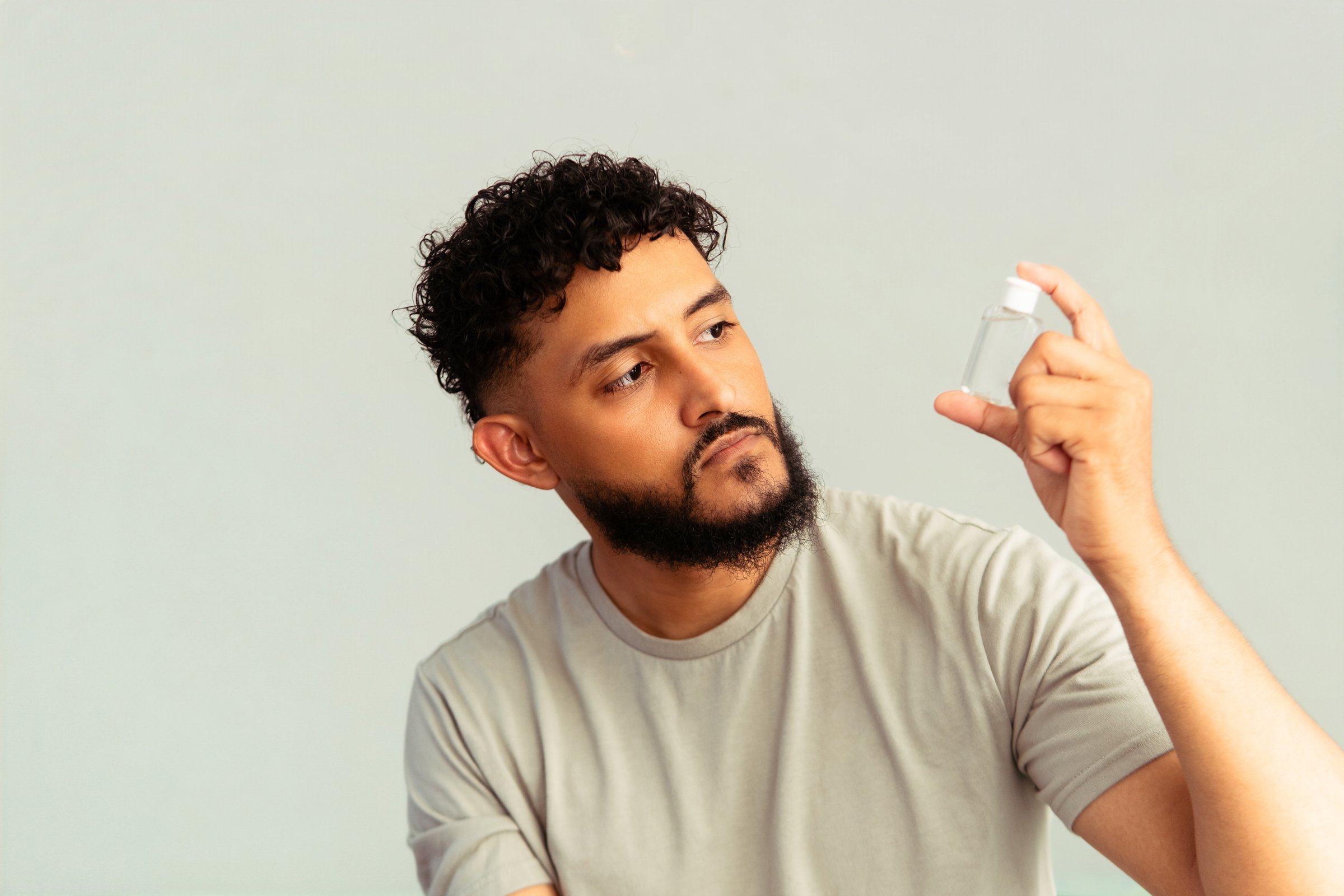 Young adult hispanic man with curly hair and beard holding a clear bottle, reviewing a skincare or hair care product