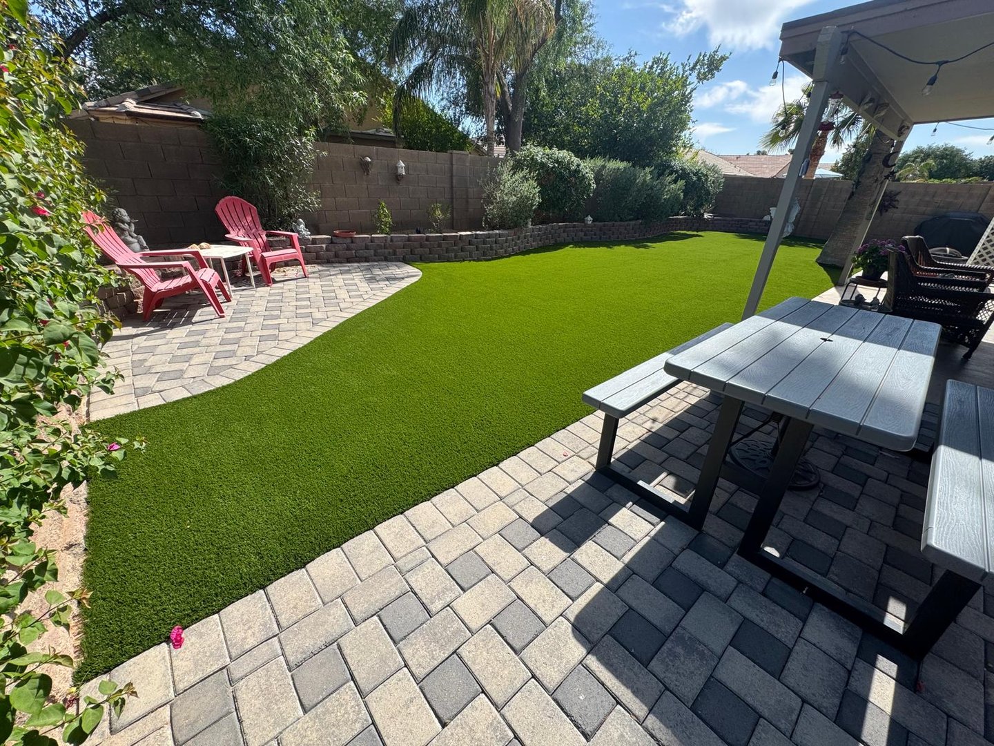 Backyard with artificial grass, patio area with red chairs, gray pavers, and a picnic table under a canopy.