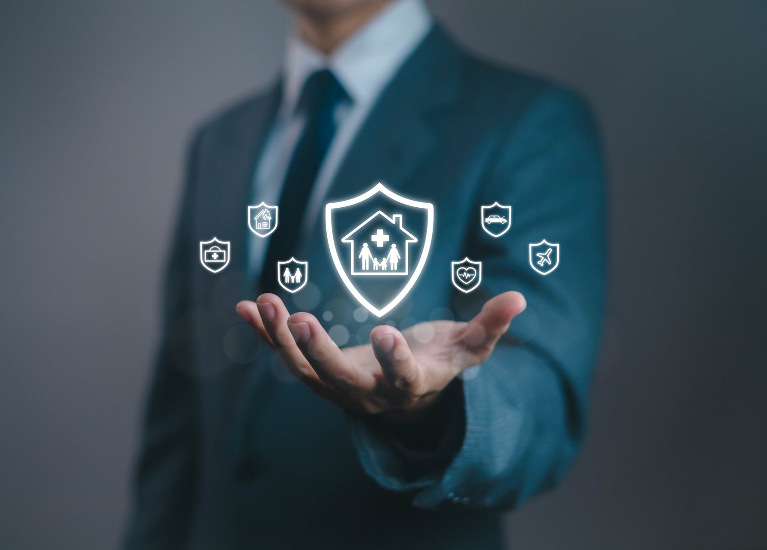 Businessman holding glowing insurance shield icon with family, health, car, and travel symbols, representing protection, security, and safety for modern financial and insurance services.