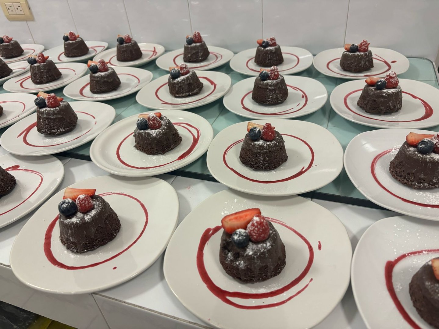 Chocolate desserts topped with berries on white plates, arranged in rows with a red sauce swirl.