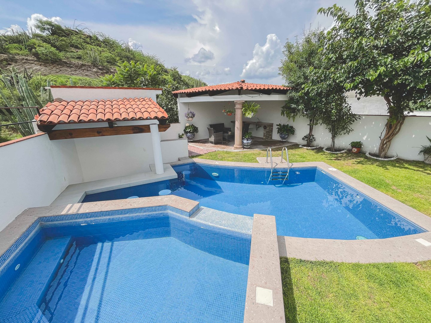 Outdoor pool area with a small hot tub, surrounding greenery, and a shaded patio with seating.