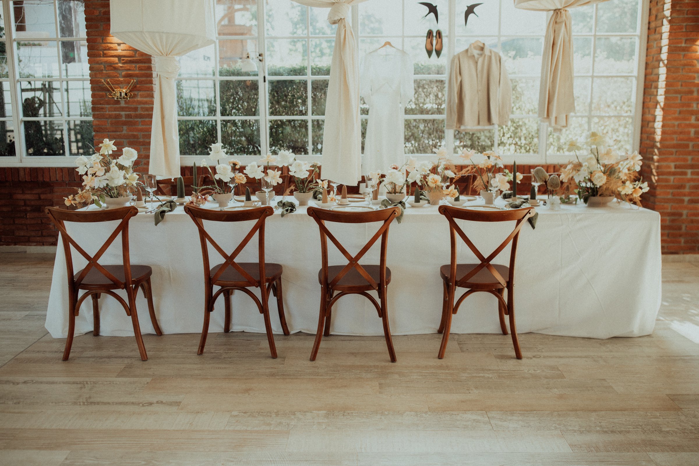 Empty chairs surrounding a wedding table with flower arrangements and the bride and groom's attire hanging in the background