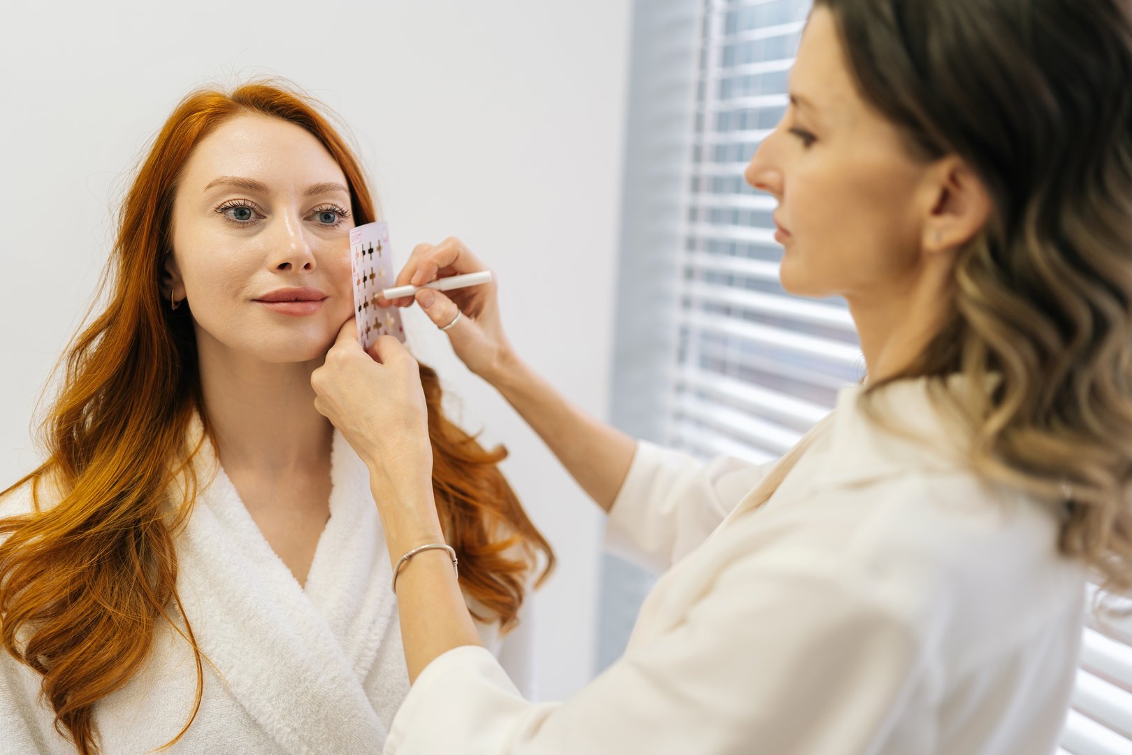 Beautician marking facial lines on redheaded client wearing bathrobe, applying transparent grid with white pen during pre-treatment skin assessment in modern aesthetic clinic