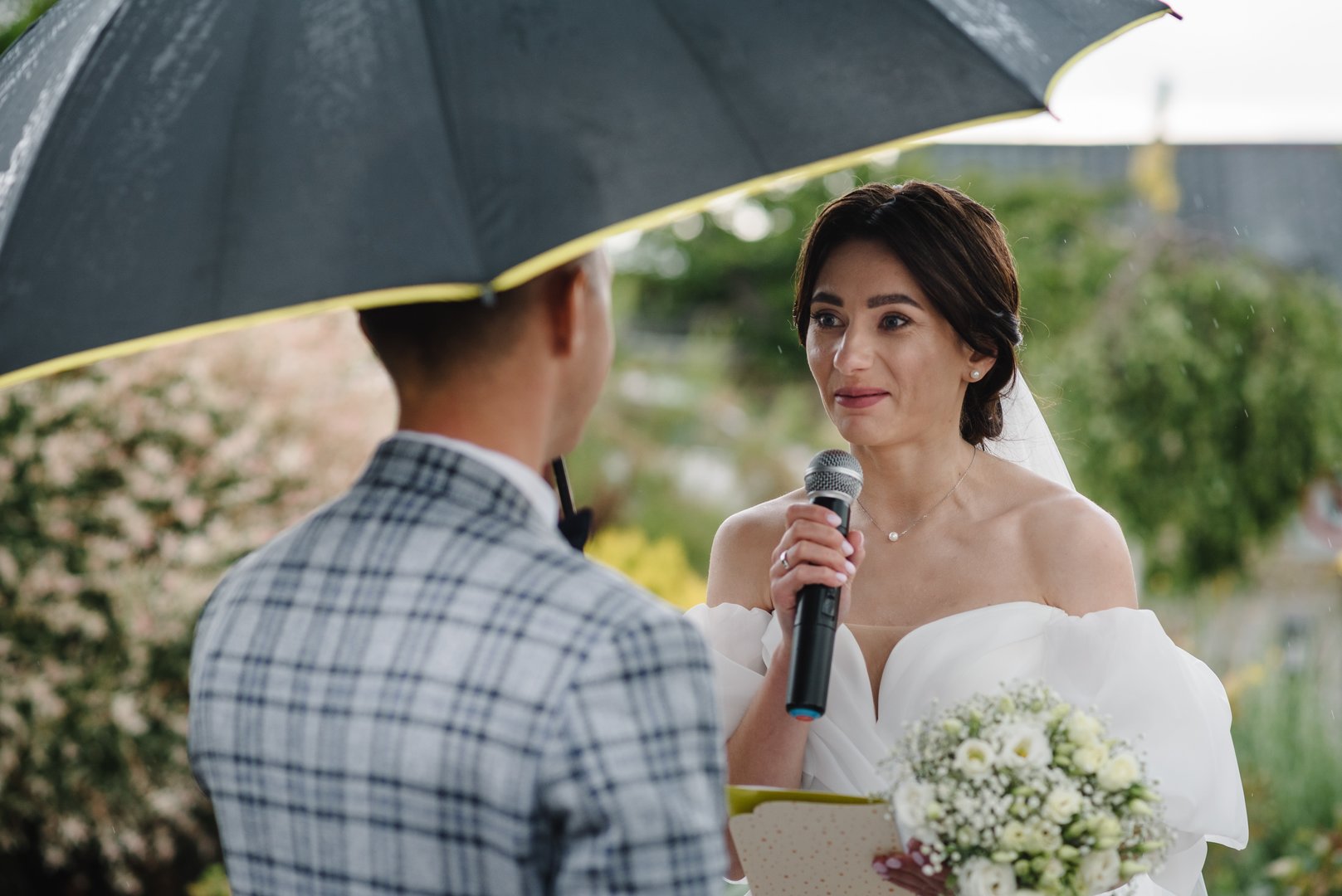Bride says promise in microphone. Happy bride and groom exchanging wedding vows on ceremony in backyard banquet area. Newlyweds standing with bouquet in rain with umbrella. Fidelity in love. Back view