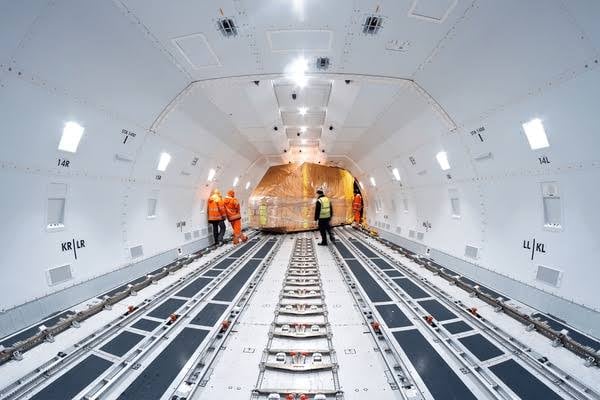 Workers in safety gear inside an empty cargo plane, supervising the loading of a large covered item.