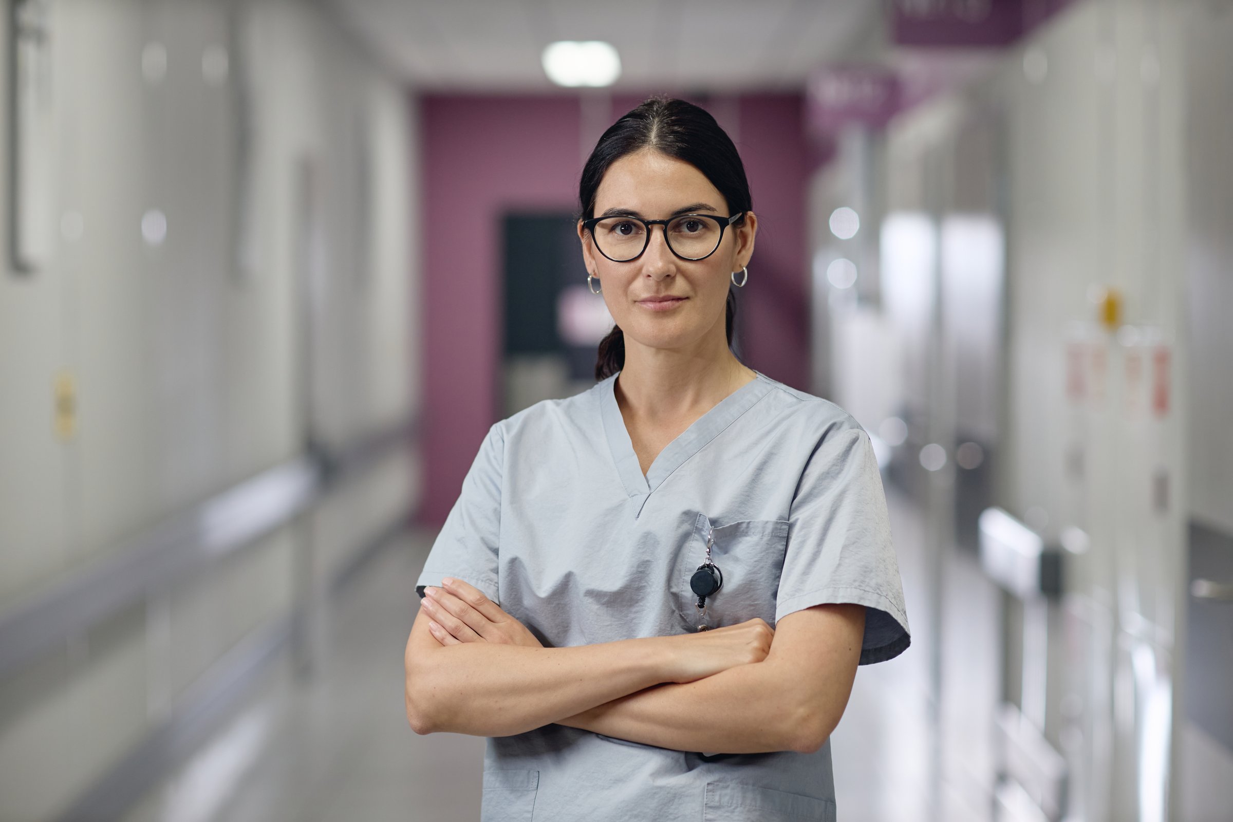 Portrait of young adult Caucasian woman standing in hospital corridor wearing medical scrubs, crossing arms and looking confidently at camera in vascular center setting