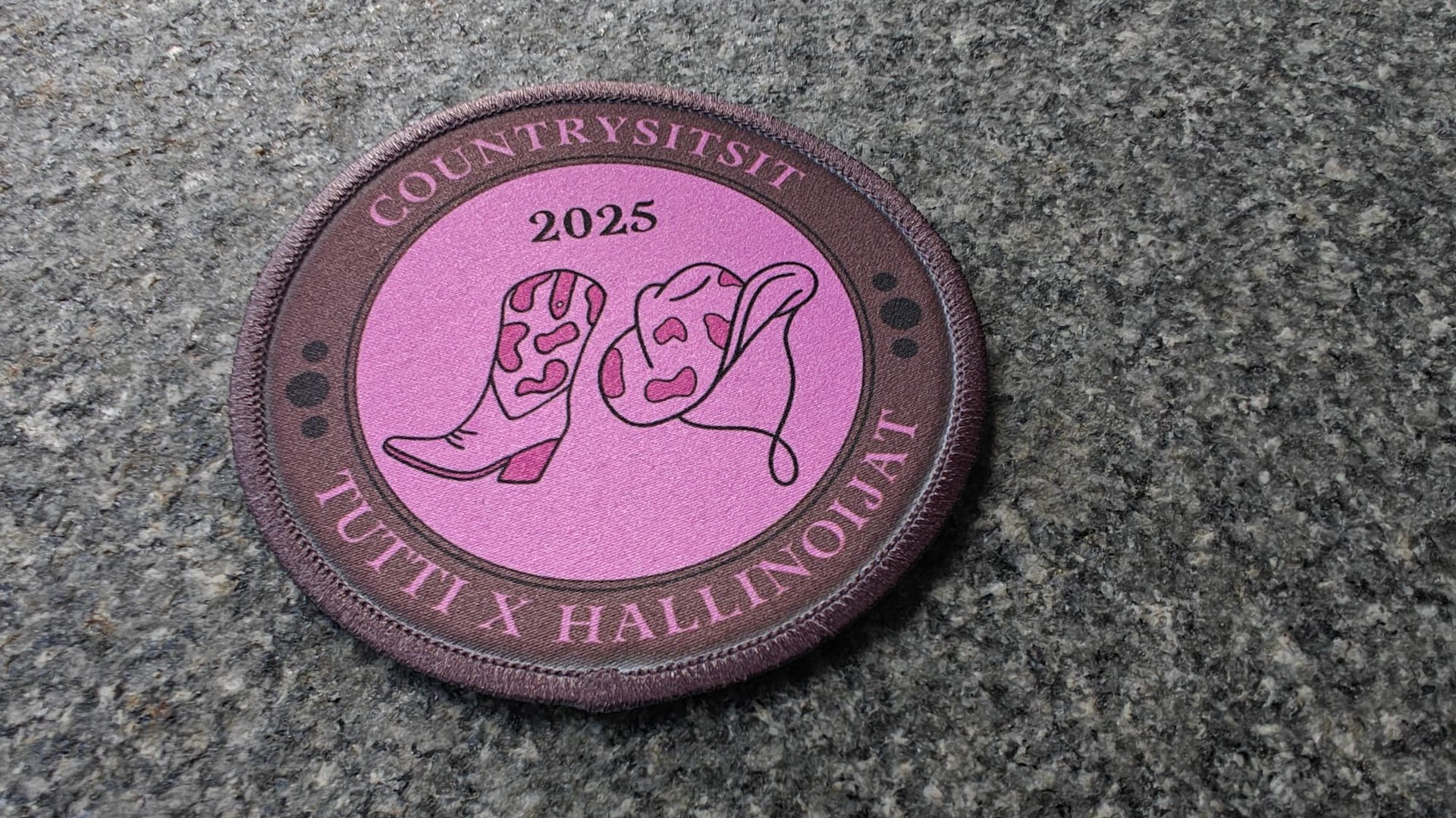 Embroidered patch with cowboy boots, text Countrysitsit 2025, and Tutti x Hallinnon, on a stone surface.