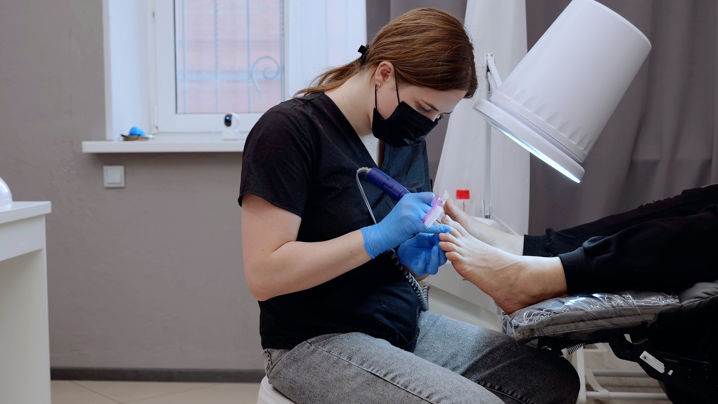 Professional hardware pedicure using electric machine in beauty salon by nail master wearing mask and gloves working on client toenails