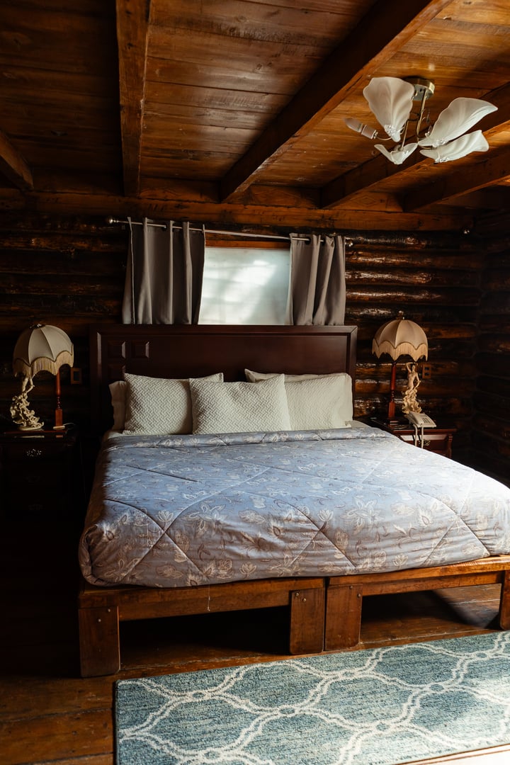 Cozy bedroom with a large bed, patterned quilt, wooden beams, curtains over the window, and vintage lamps on side tables.