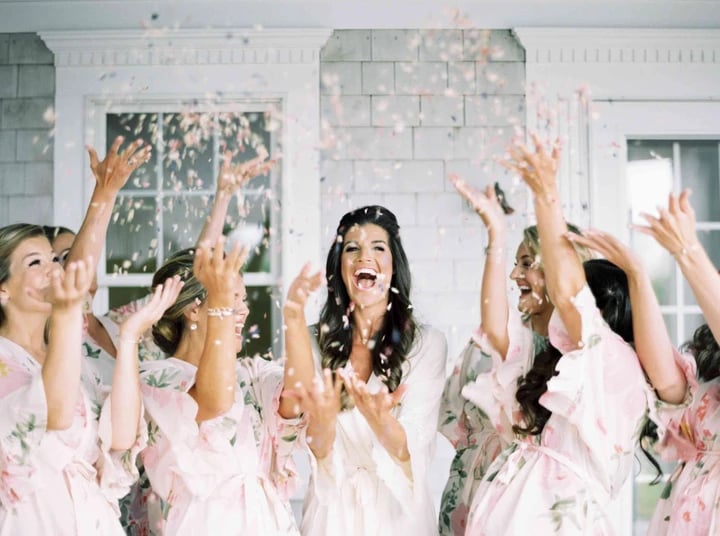 Bride and bridesmaids in robes joyfully tossing confetti indoors, celebrating with laughter and excitement.