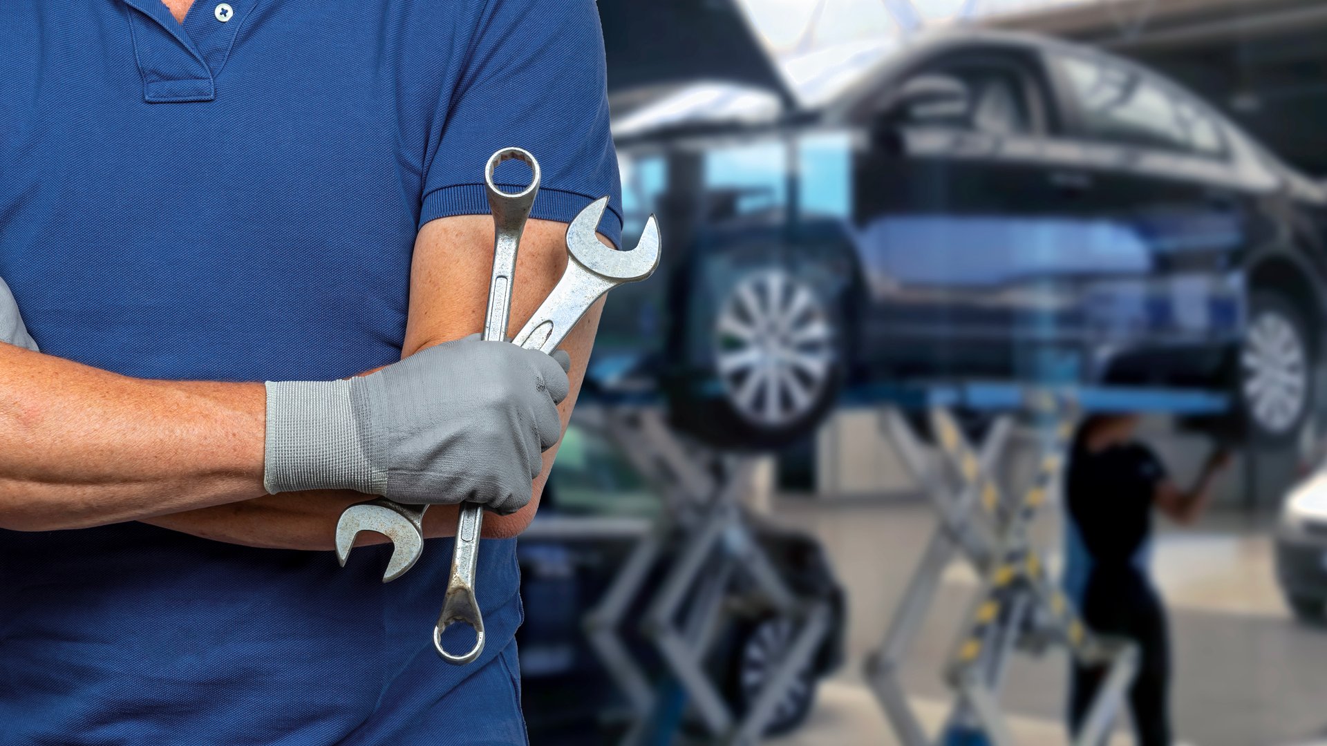 Mechanic with crossed arms and holding wrenches with a car on a lift in auto repair workshop.