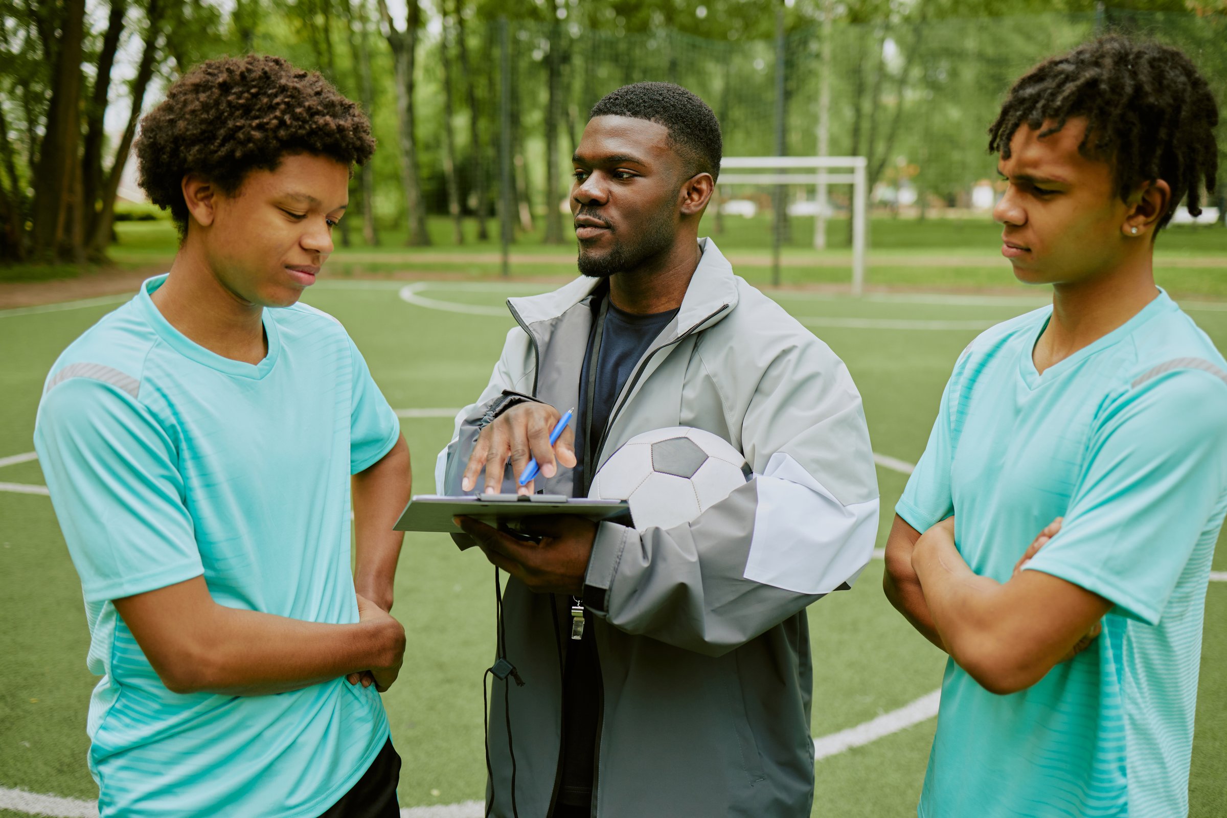 Black man holding soccer ball and clipboard instructing two teenage boys on outdoor sports field, teenagers listening attentively with arms crossed during training session