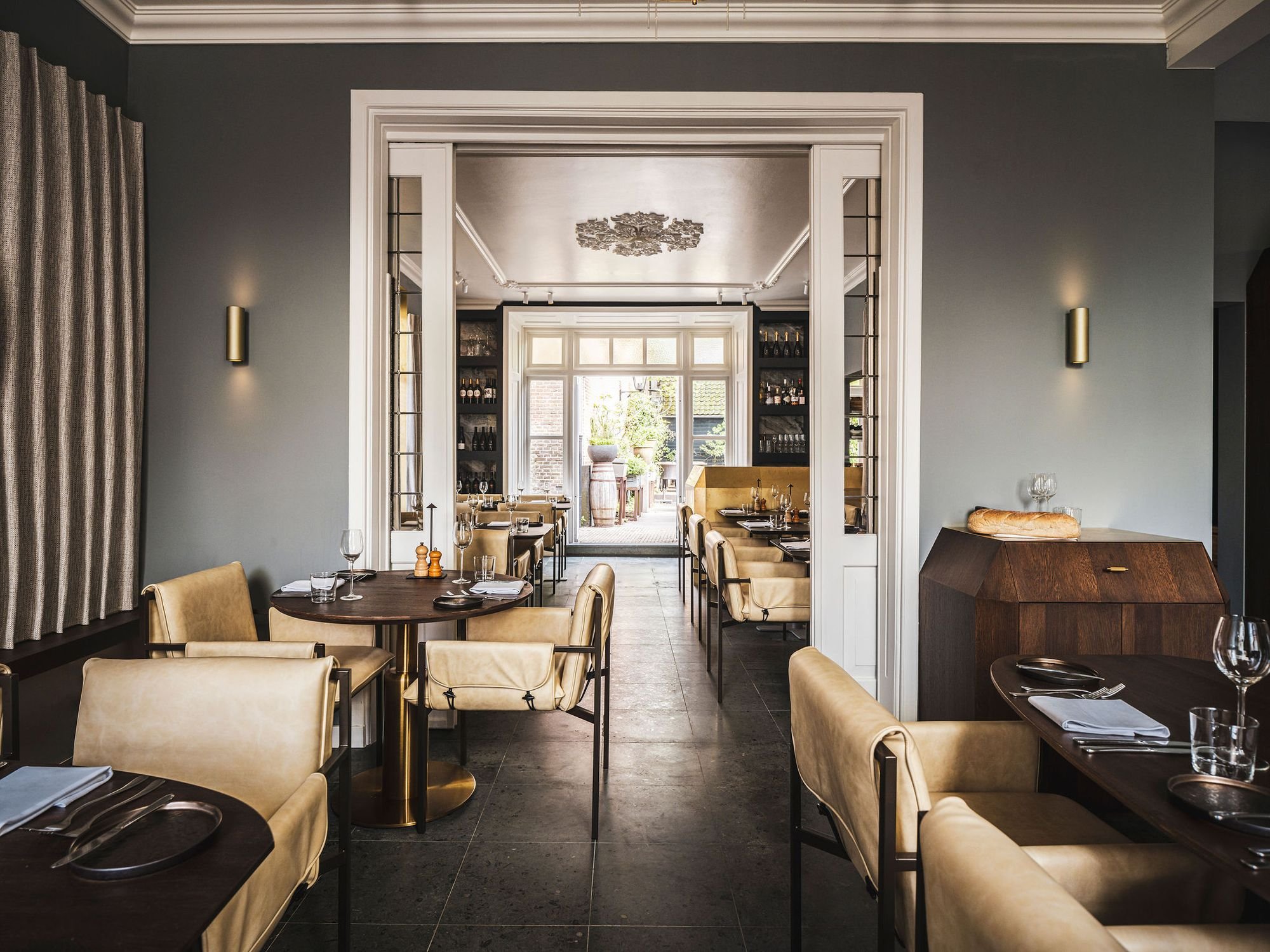 Elegant restaurant interior with beige chairs, dark round tables, and a view into a brightly lit dining area through double doors.