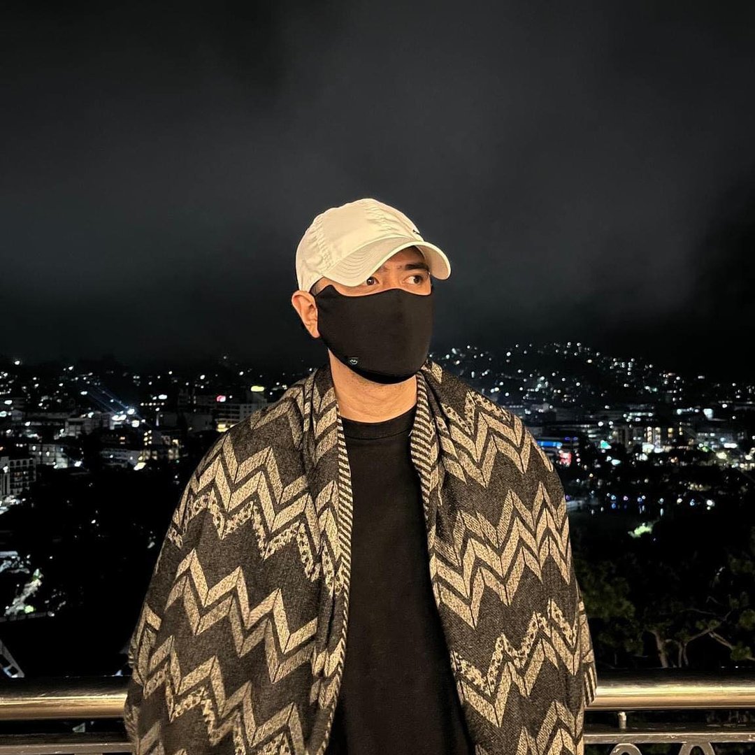 Person wearing a white cap, black mask, and patterned shawl with a cityscape in the background at night.
