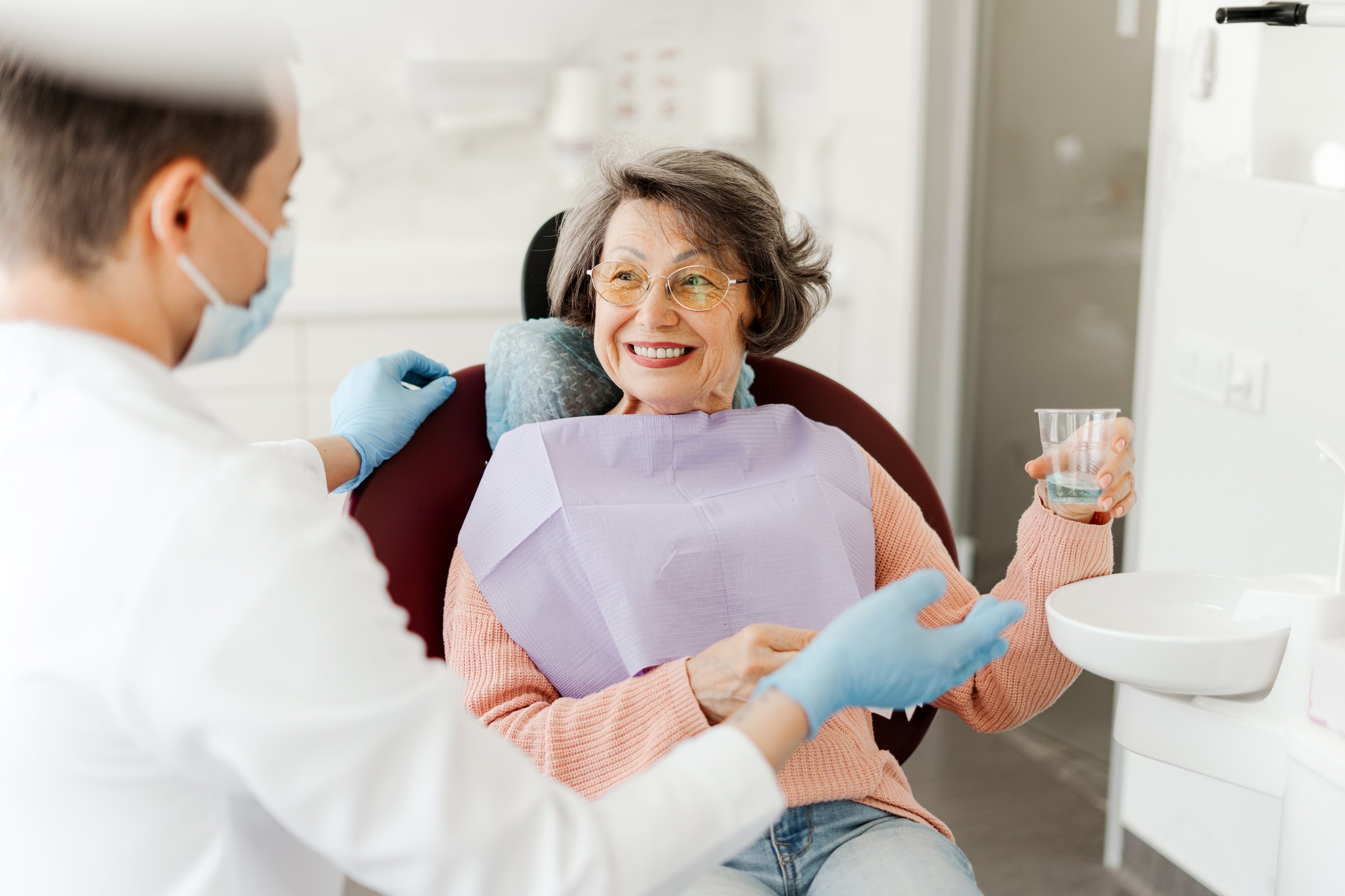 Happy beautiful senior woman sitting in dental chair, holding glass of water, talking with dentist doctor after dental treatment. Concept of service, dental care