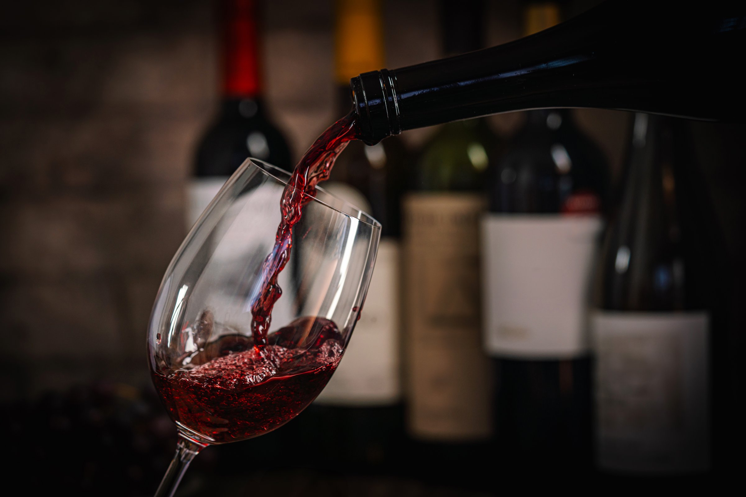 A dark, elegant still life of red wine being poured into a glass with wine bottles and fresh grapes in the background. Perfect for wine-themed compositions