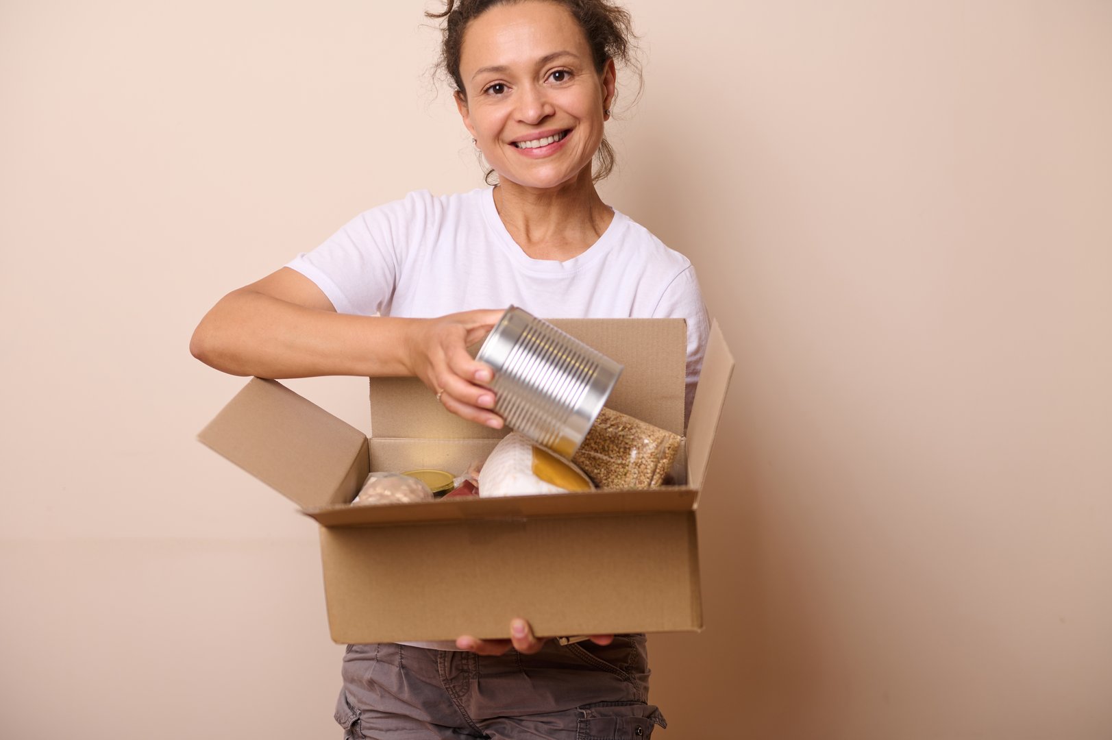 Isolated on beige studio background smiling volunteer woman wearing white t-shirt, packing goods into mockup cardboard box, preparing humanitarian help for charity and donation. Food Bank Day
