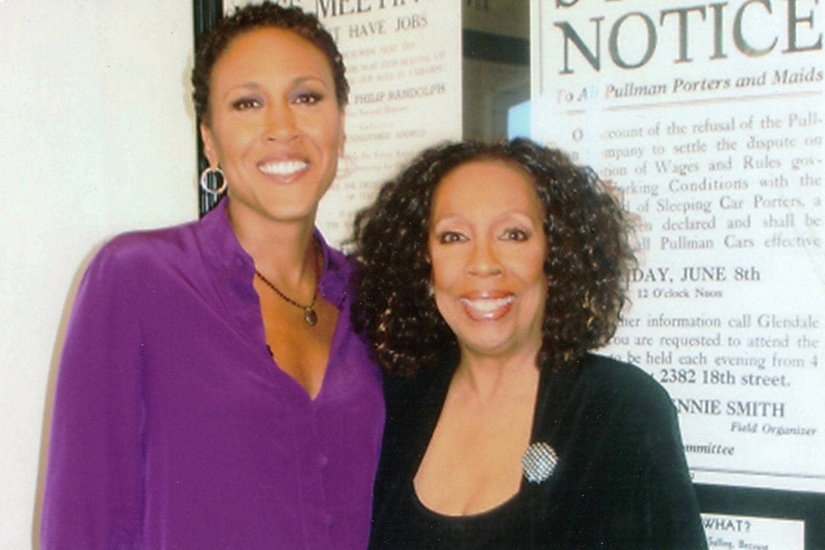 Two women smiling, one in a purple blouse, the other in black, standing indoors in front of posters.