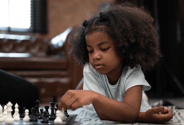 Young girl with curly hair playing chess on a board, focused on moving a piece, sitting on a patterned rug indoors.