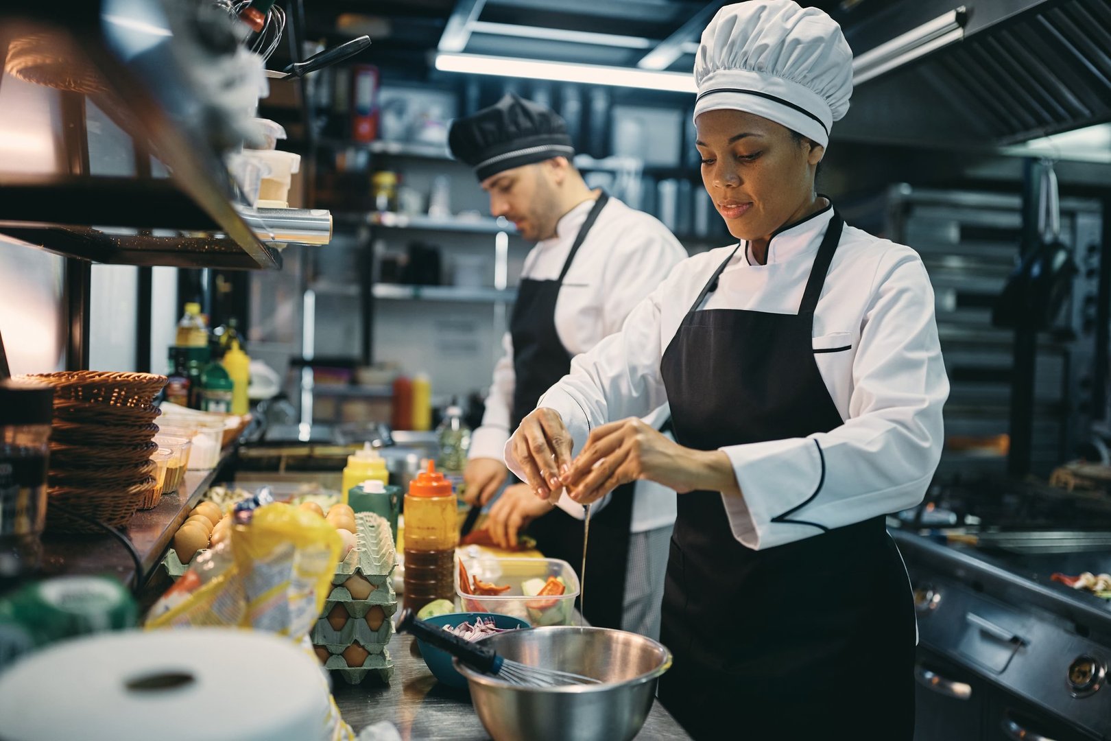 African American professional cook cracking an egg while preparing meal with her coworker in the kitchen at restaurant.