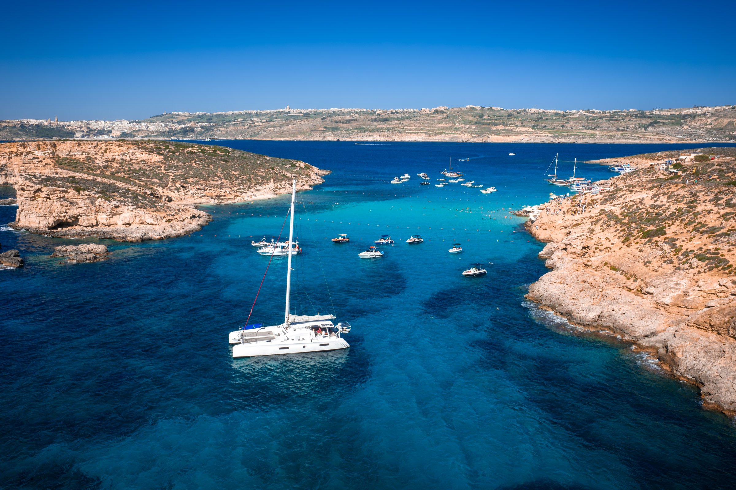 Catamarans and small boats drift on turquoise water at Blue Lagoon, Comino Island, Malta. Aerial wide angle, high saturation, clear day, rocky cliffs, narrow channel.