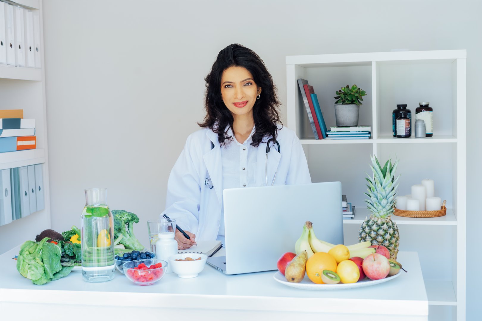 Positive Indian female nutritionist having online consultation with client at office desk with fresh vegetables, prescribing nutritional supplements and vitamins, making balanced corrective diet plan