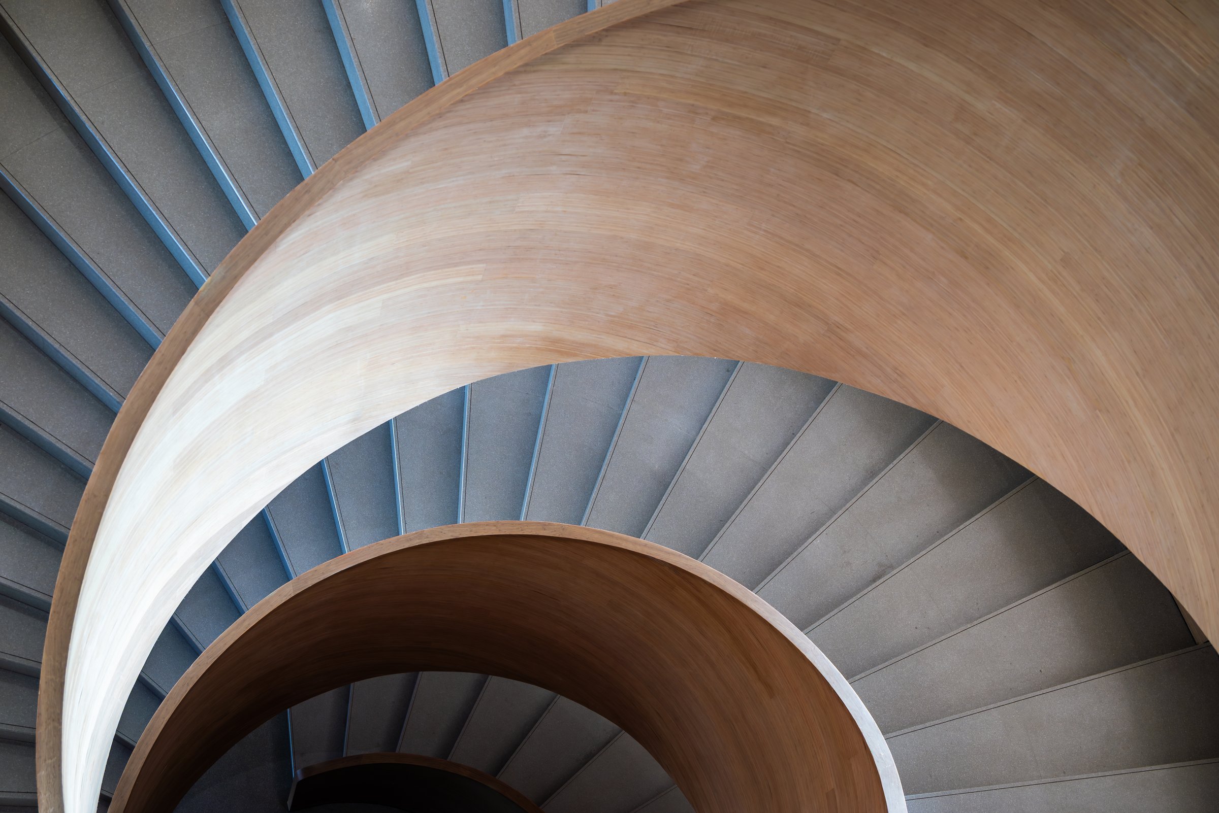 Modern spiral staircase view from above. The staircase is constructed with smooth, light-colored wood, creating a sleek and elegant appearance. The steps are made of concrete, adding a contrasting texture to the wooden surface. The design is minimalist, with clean lines and a curved structure that adds a sense of movement and flow. The staircase is well-lit, with subtle lighting highlighting the texture of the wood and the steps.