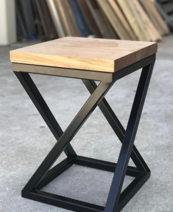 A modern stool with a wooden top and a black metal geometric frame, set against a background of stacked wooden boards.
