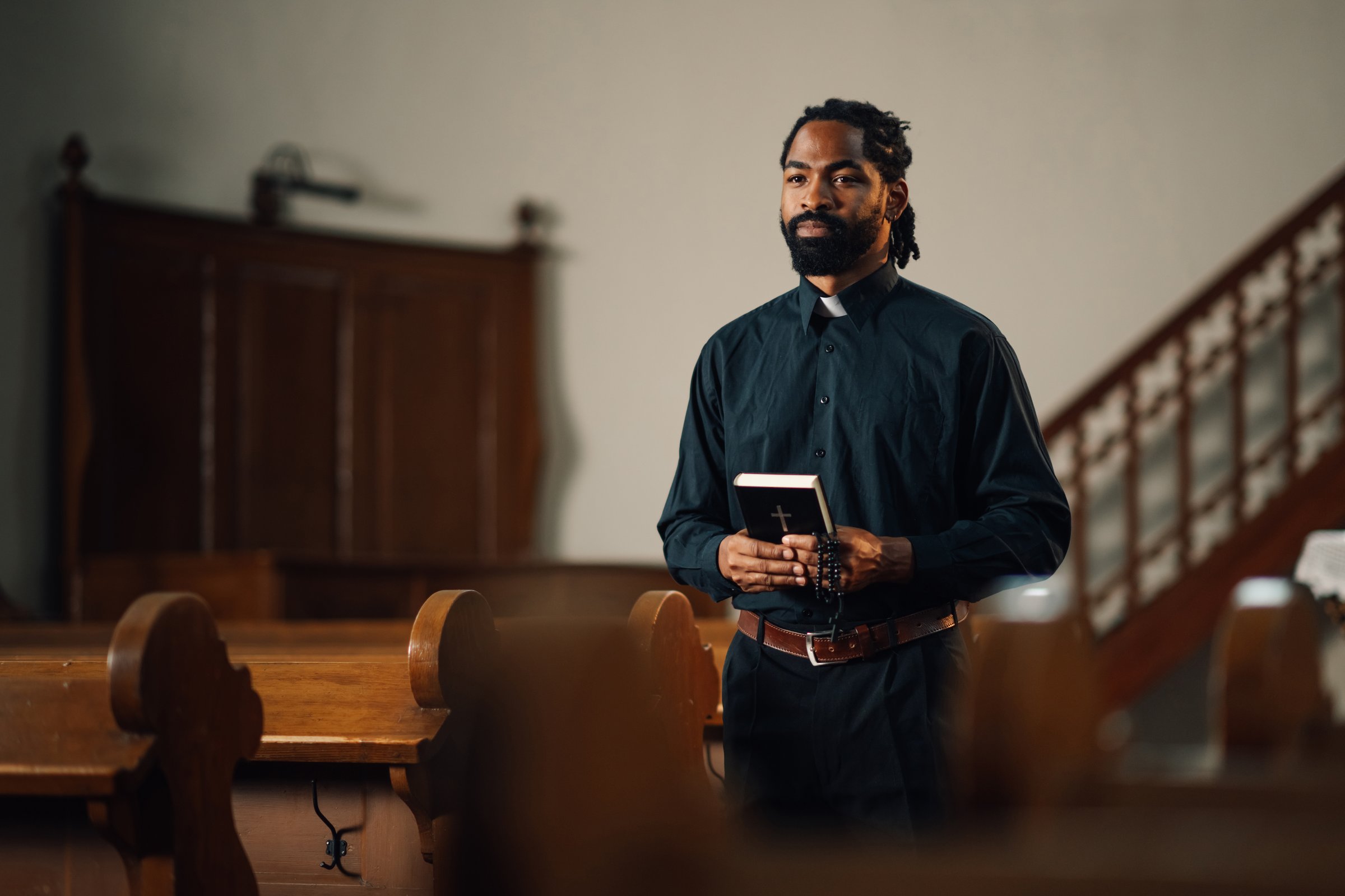 Priest is holding a bible and rosary beads while standing in a church. The image evokes faith, spirituality and religion