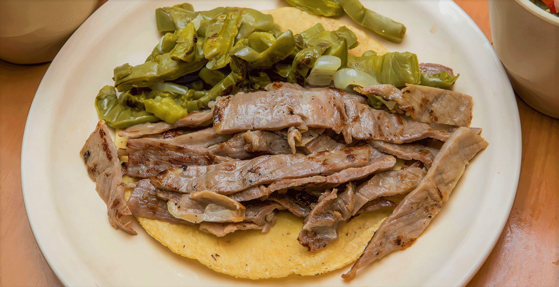 Grilled meat slices and cooked green peppers on a tortilla, served on a white plate.
