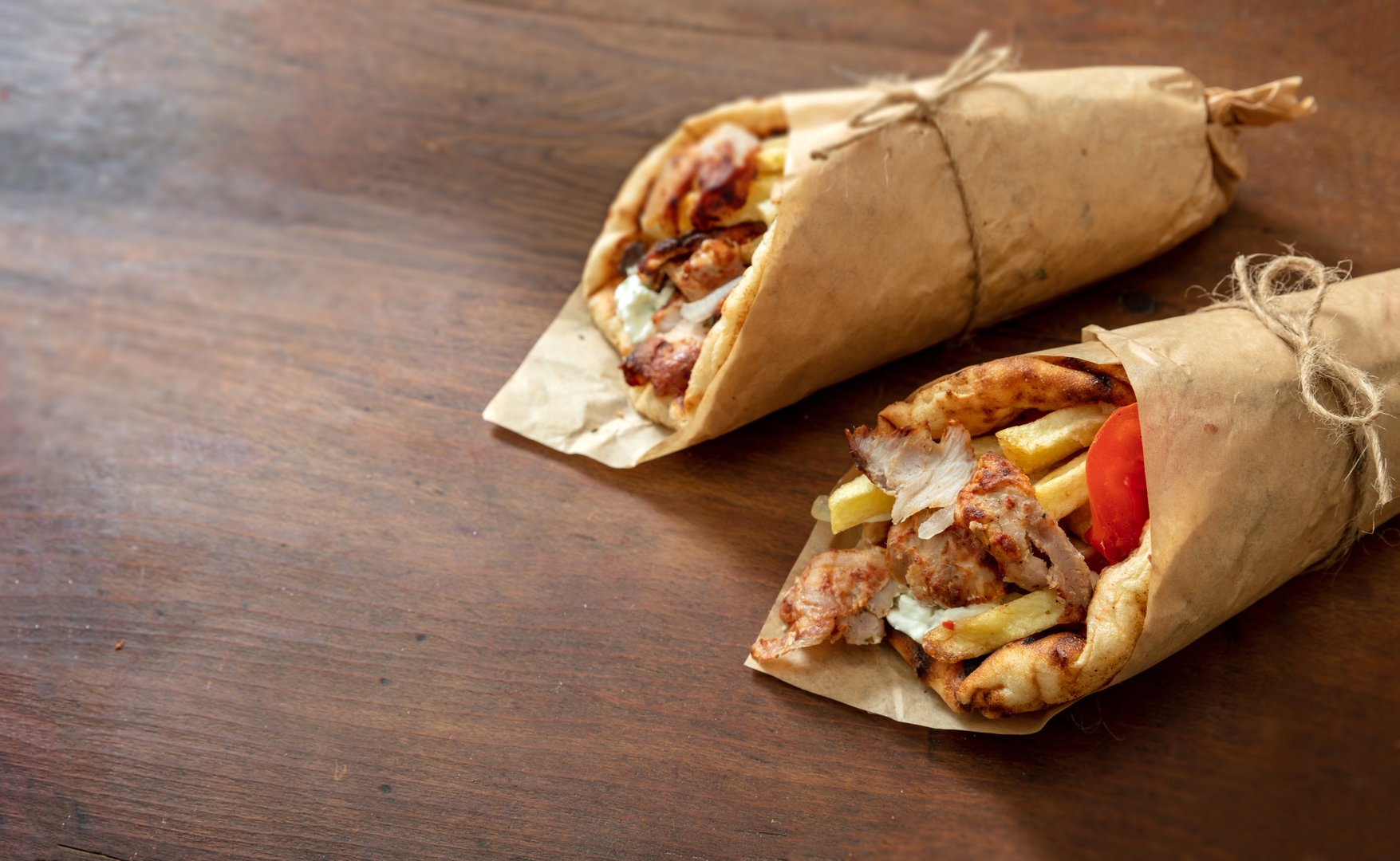 Two freshly wrapped chicken gyros with fries, tomato, and sauce rest on a rustic wooden table in warm natural light