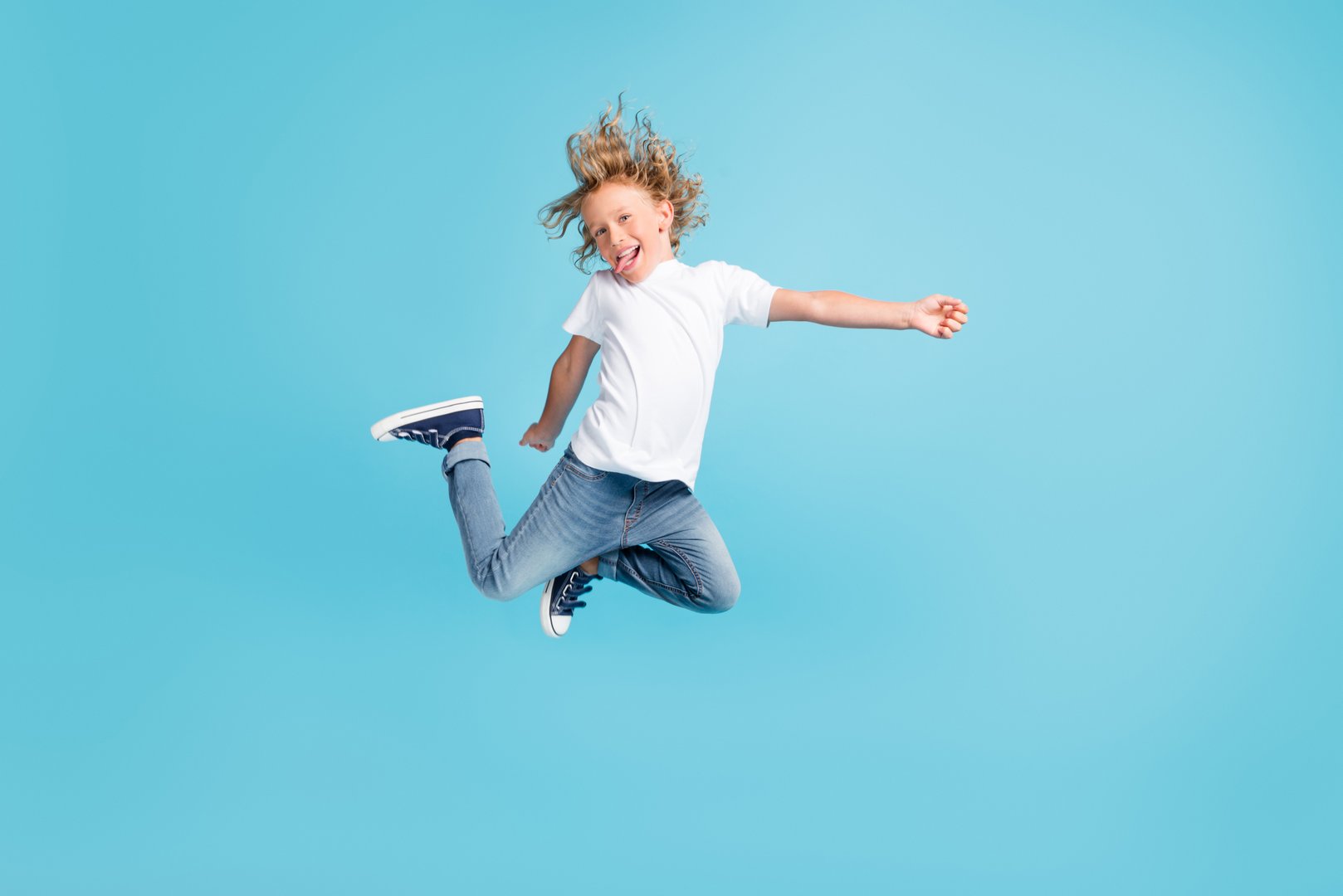 Photo of little child jump raise legs stick-out tongue wear white shirt jeans, sneakers isolated blue color background