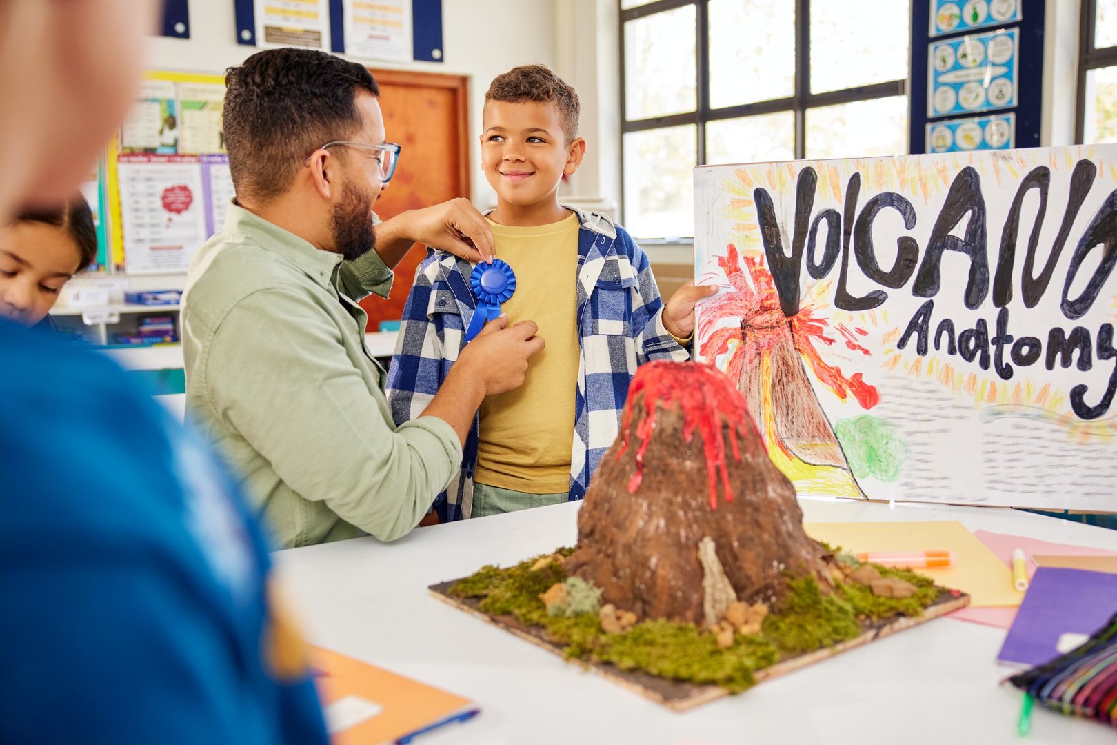 Teacher presenting award badge to schoolboy for volcano science project at elementary school