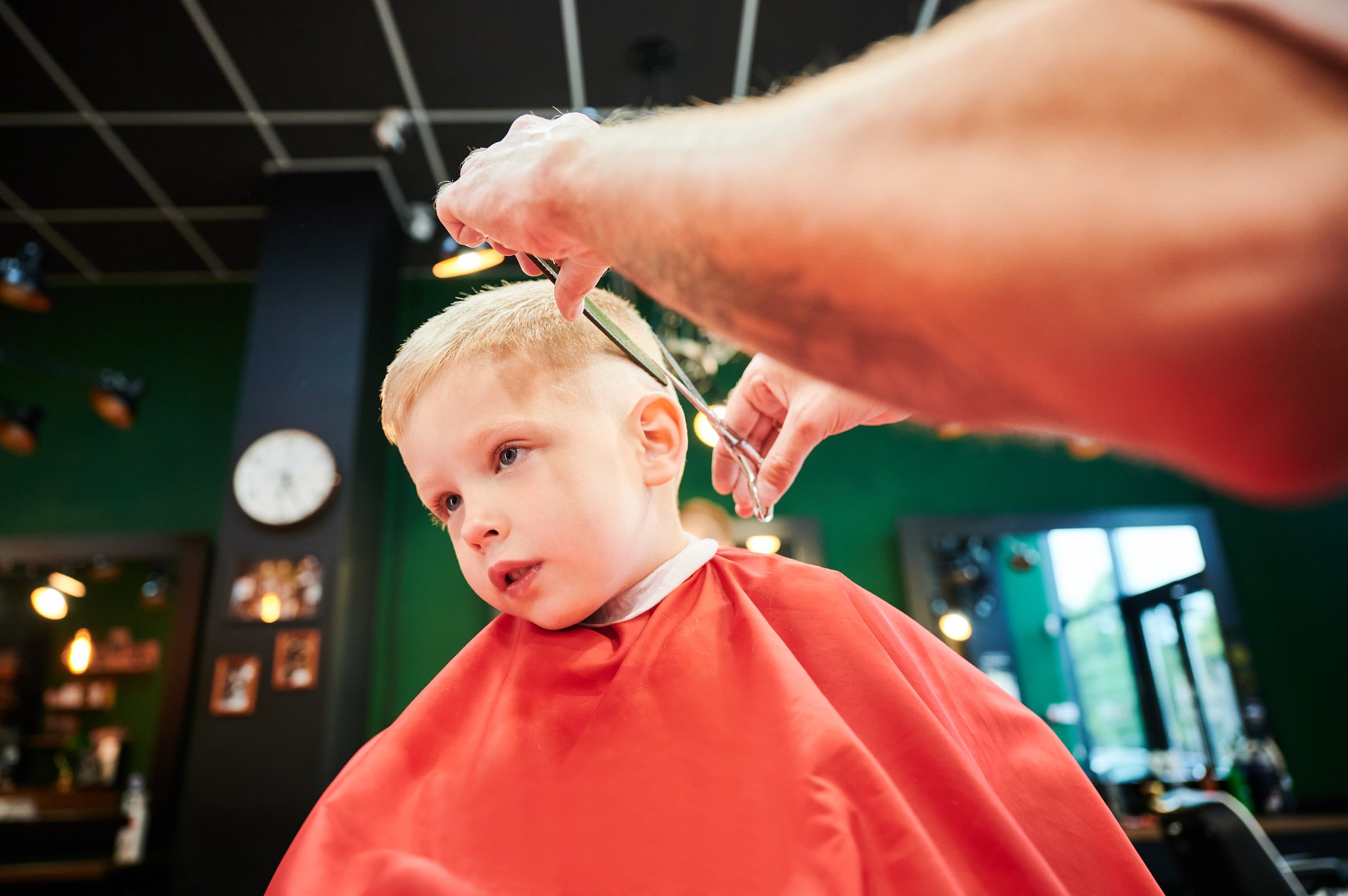 Man barber cutting little boy's hair using comb and scissors. Child getting haircut from adult male, likely barber. Professional hairdresser and cute client at modern barbershop.