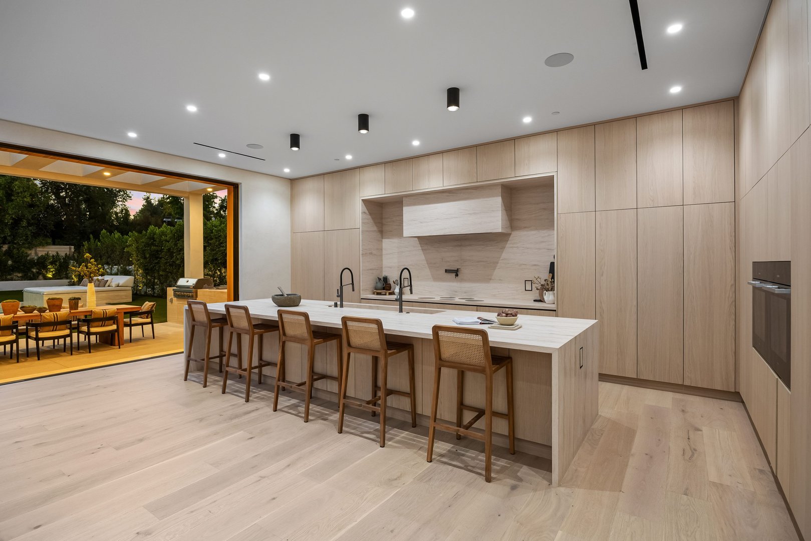 Modern kitchen interior with wooden cabinets and island, open to outdoor dining area, featuring sleek design and natural light.