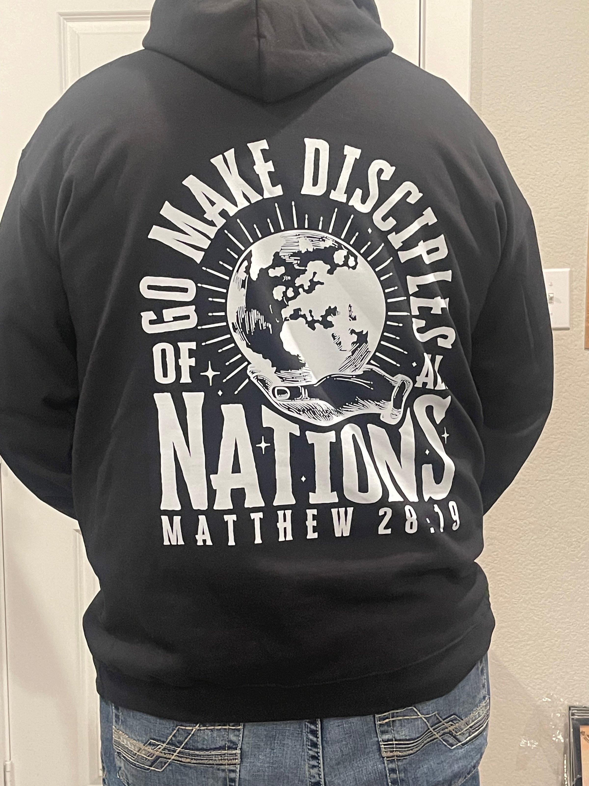 Person wearing a black hoodie with text: Go Make Disciples of All Nations Matthew 28:19, featuring a globe and sunburst design.
