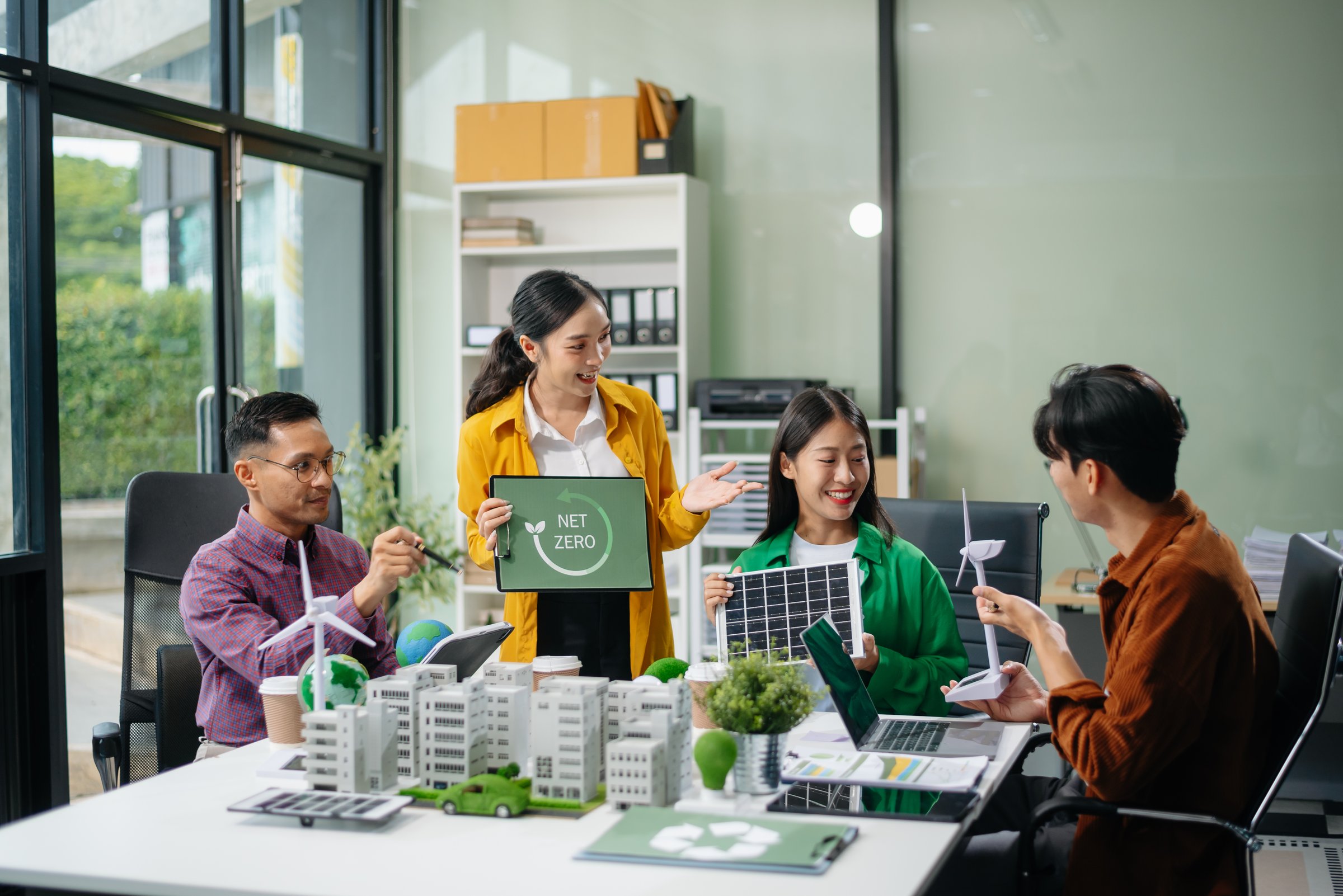 Asian professionals collaborate on sustainable energy solutions with wind turbines, solar panels, and eco friendly models in office
